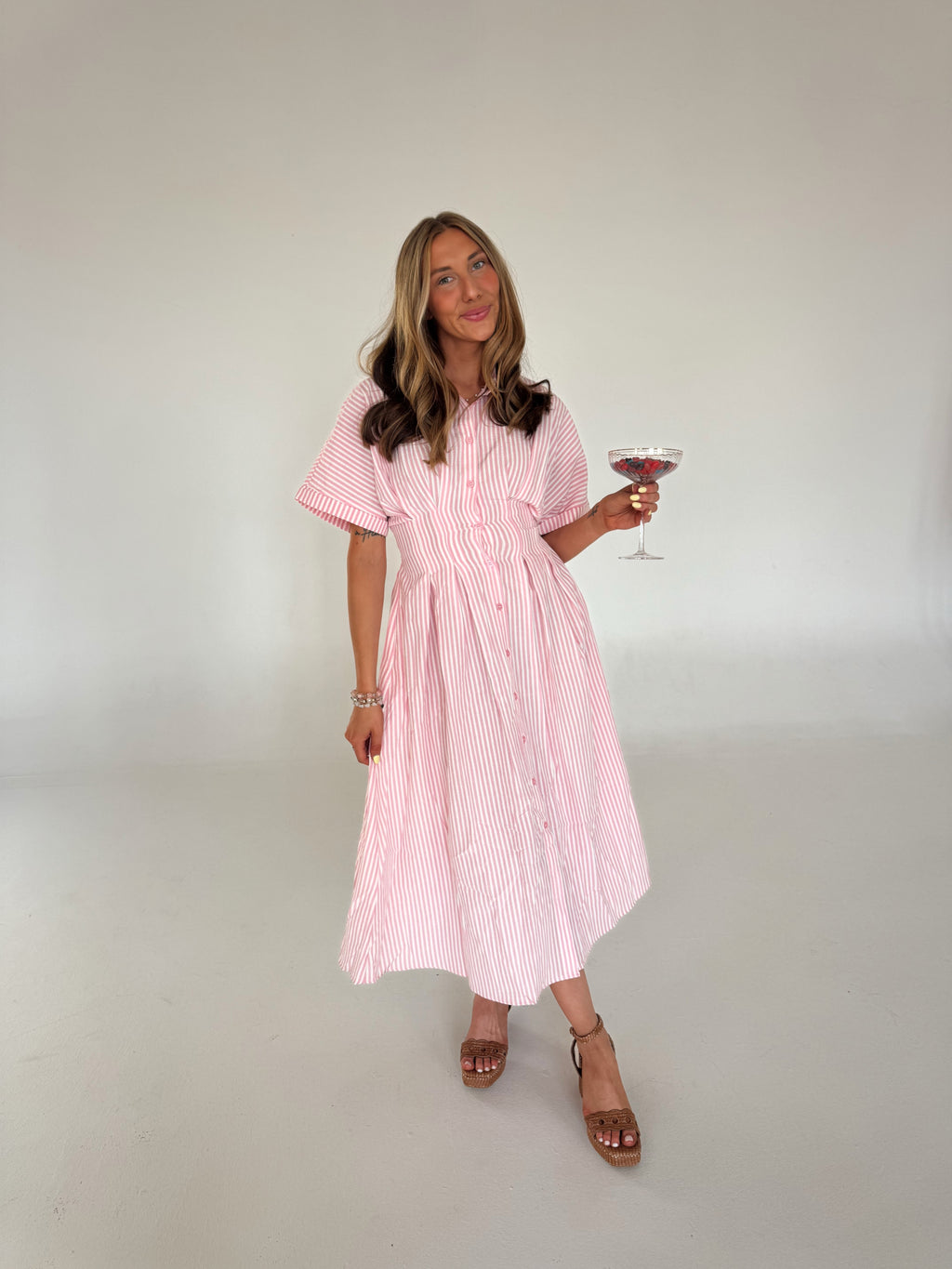 Woman in a pink and white striped dress holding a glass of red wine against a plain background