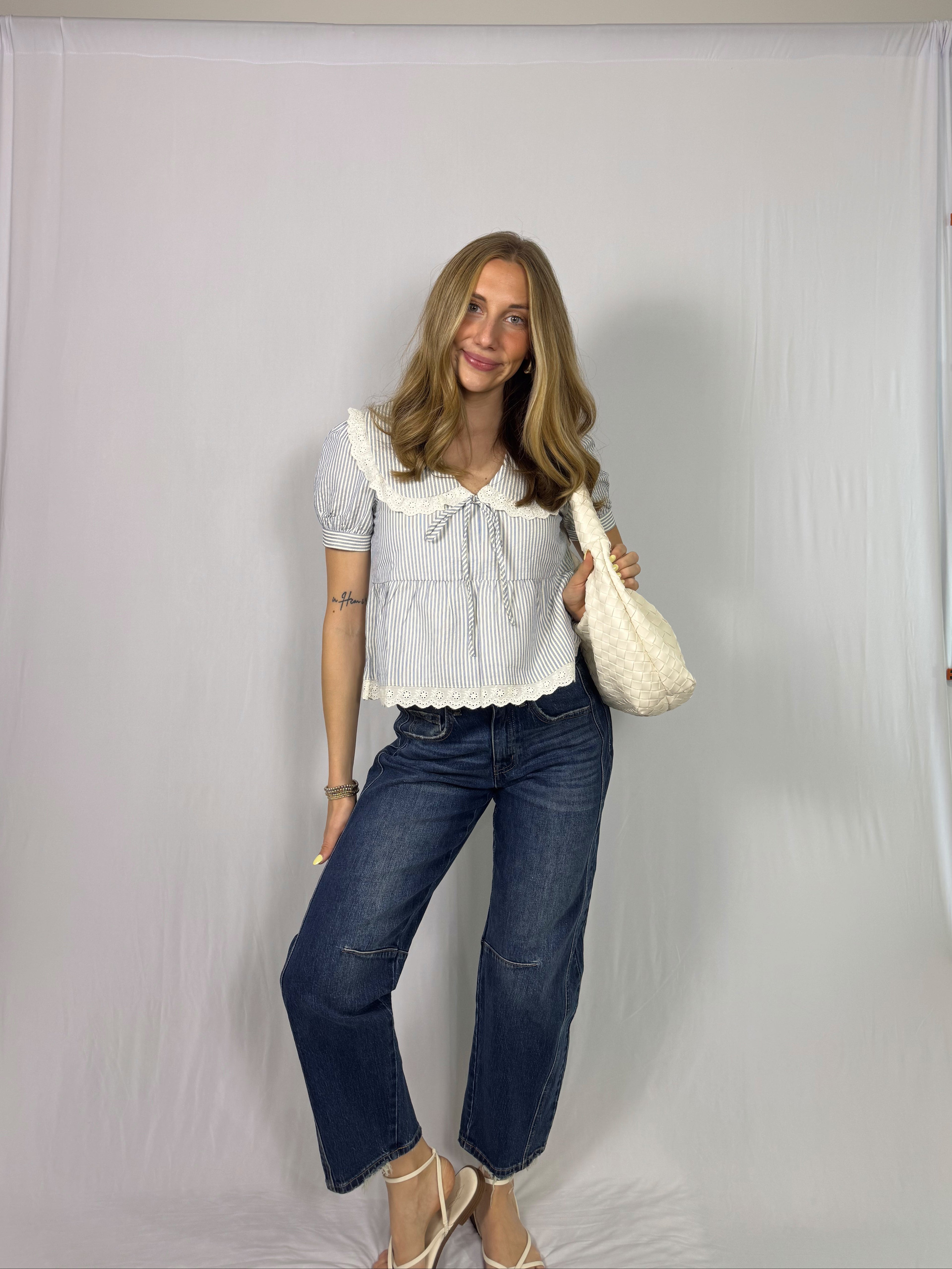 Woman wearing a striped blouse with ruffled collar and blue jeans, holding a cream-colored bag against a plain background.