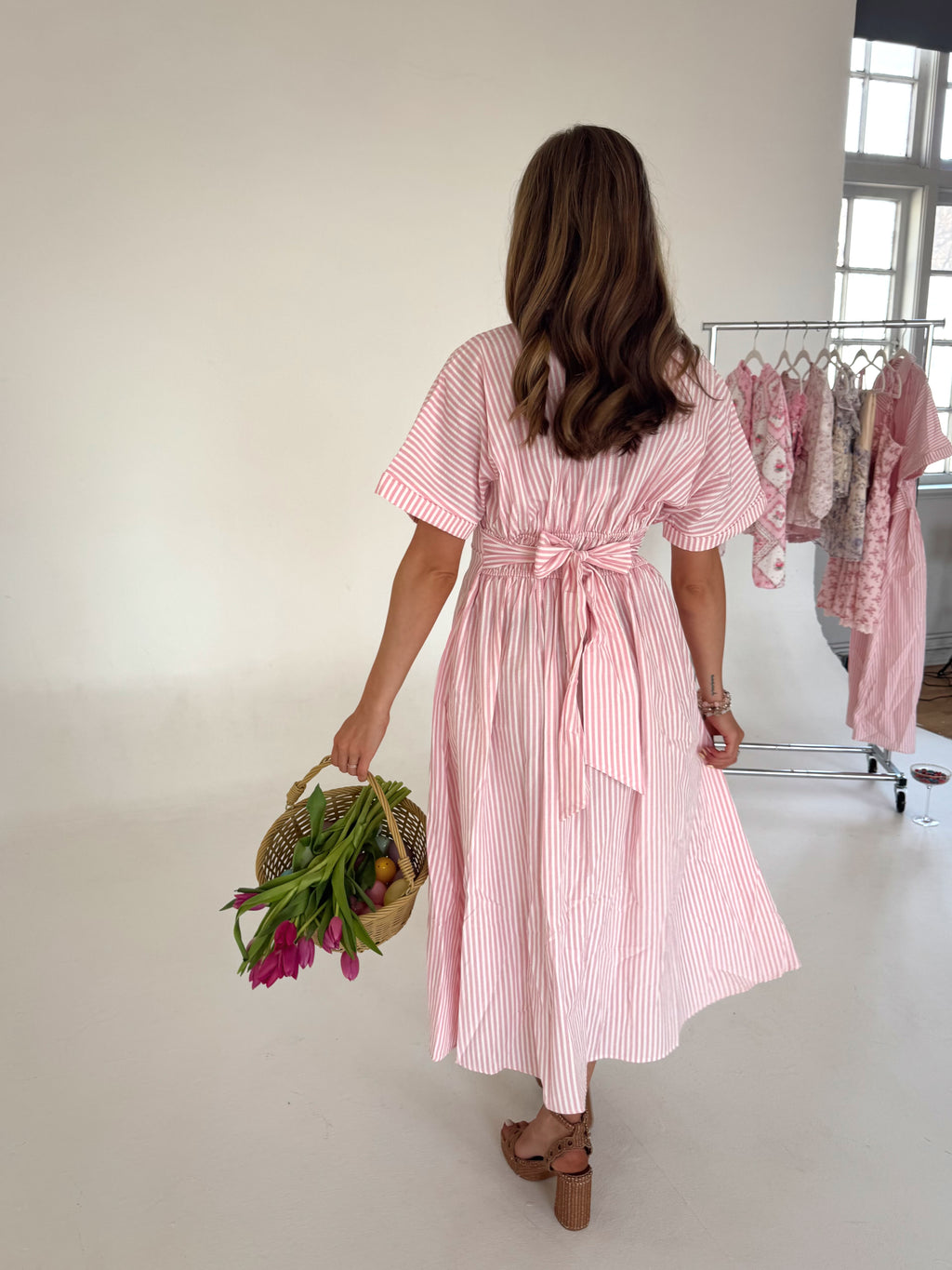 Woman in a pink striped dress holding flowers in a room with clothing racks.