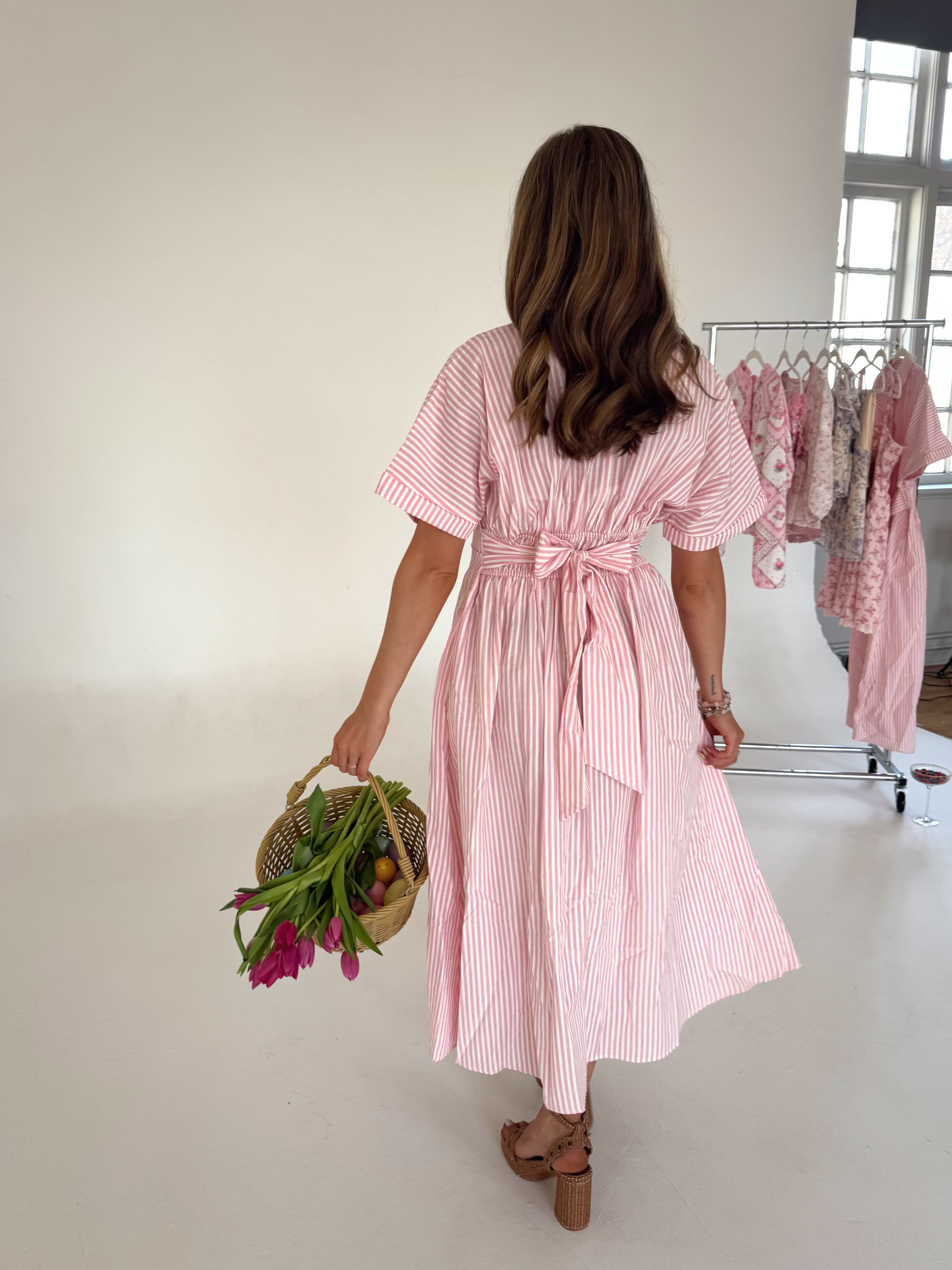 Woman in a pink striped dress holding flowers in a room with clothing racks.
