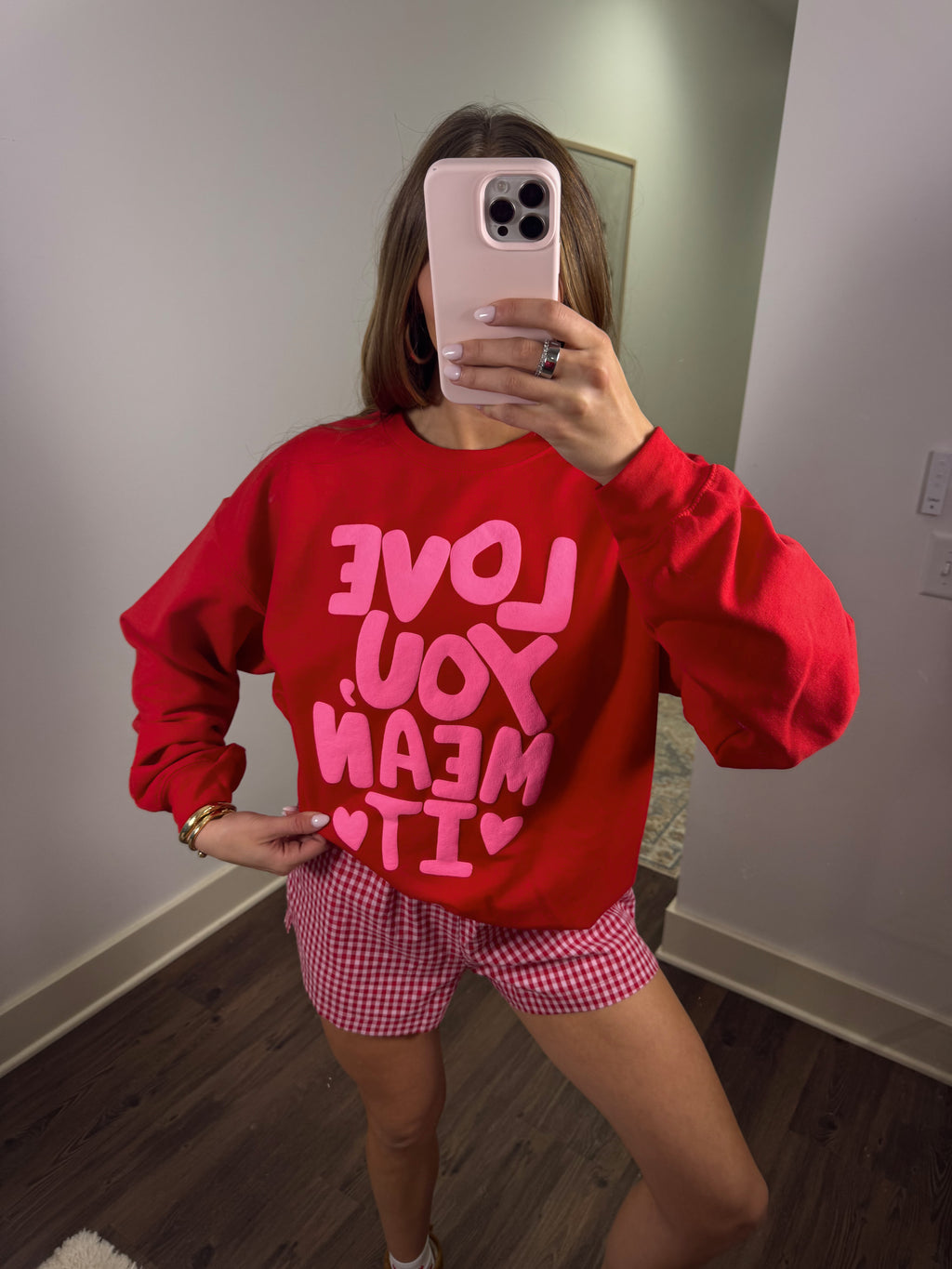 Person wearing a red sweatshirt with pink text and pink checkered shorts in a room.