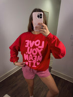 Person wearing a red sweatshirt with pink text and pink checkered shorts in a room.