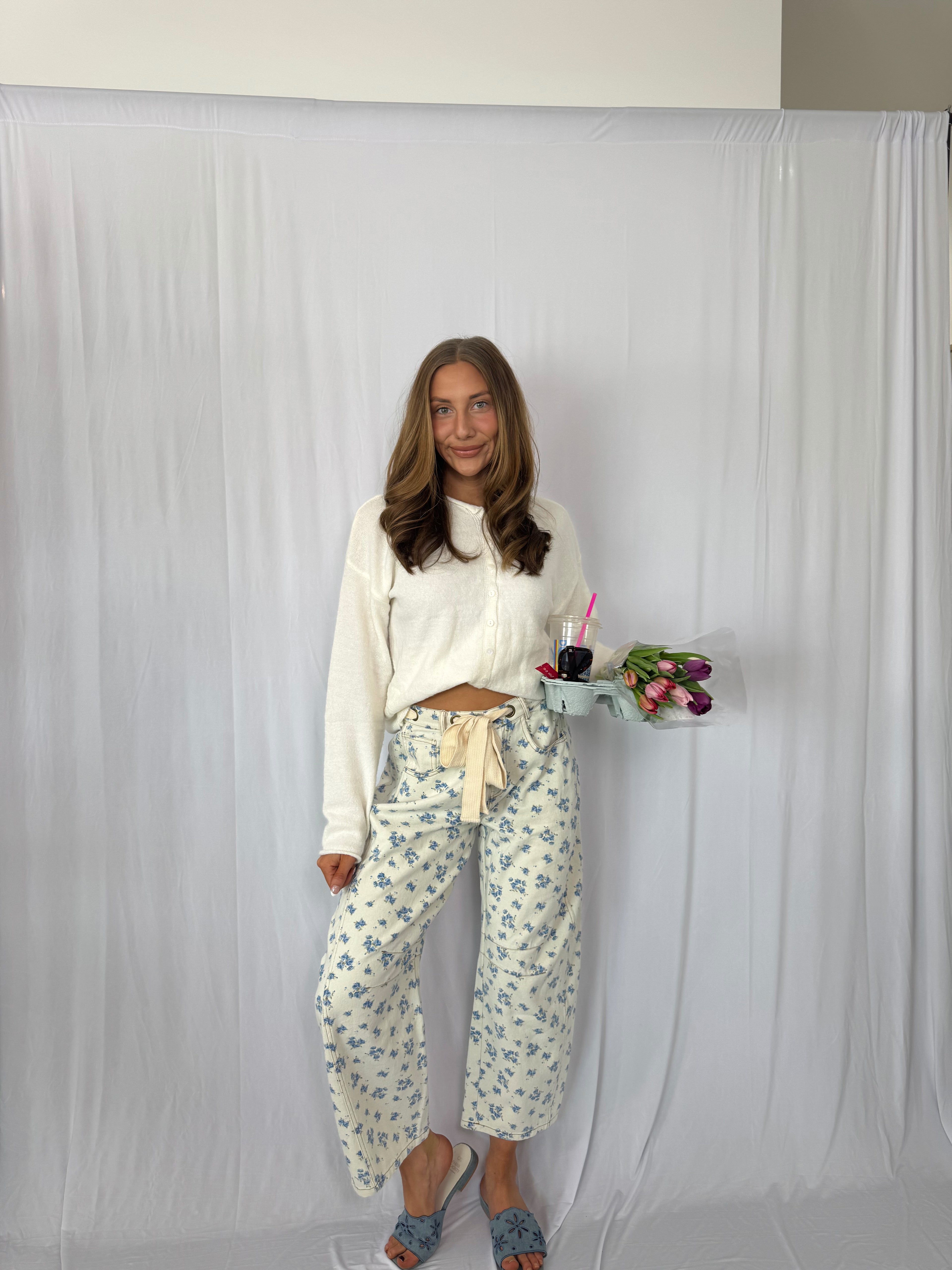 Woman in a white top and floral pants standing against a plain background with a tray of drinks and flowers.