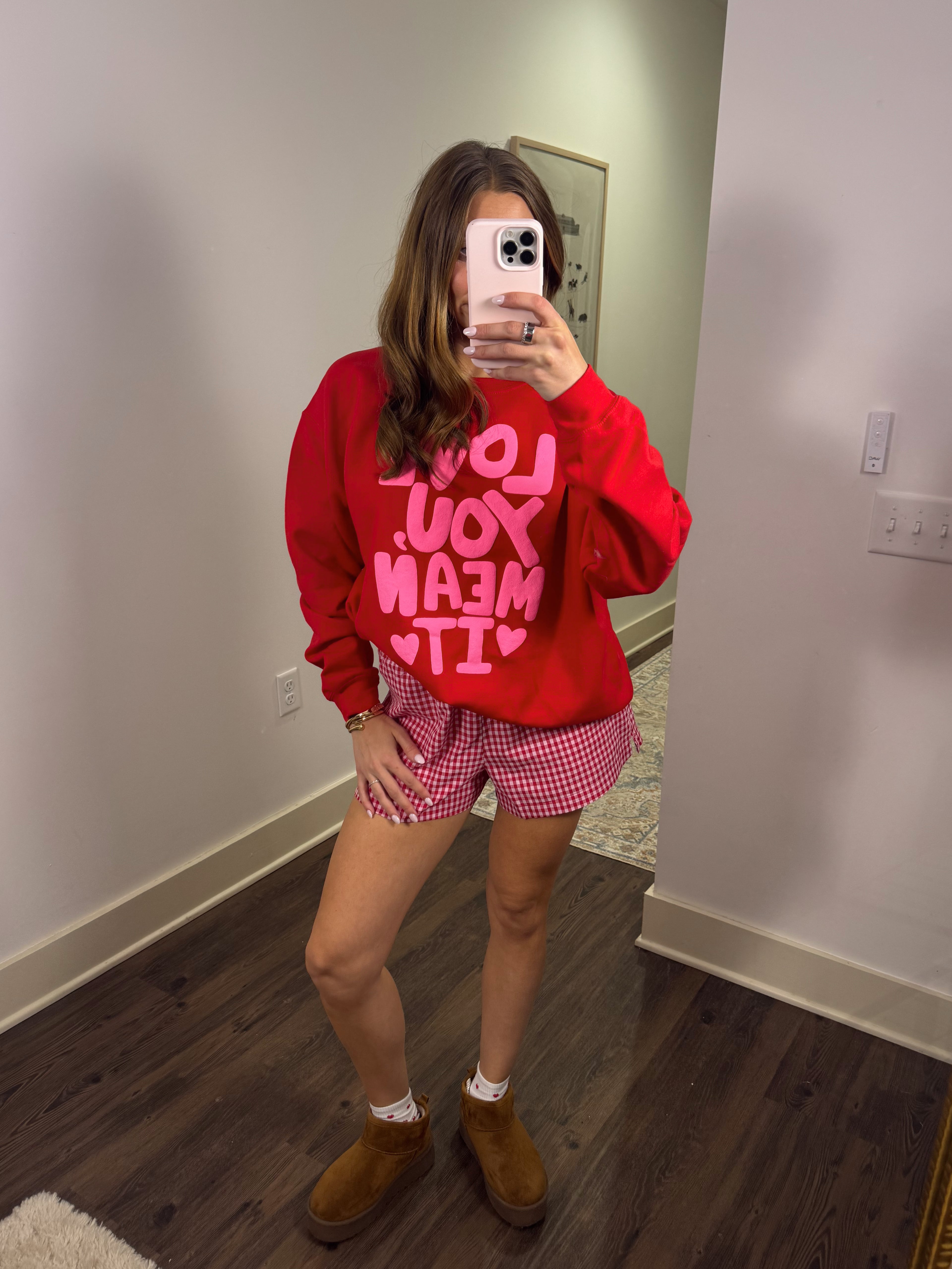Person wearing a red sweatshirt with pink text and pink checkered shorts in a room.
