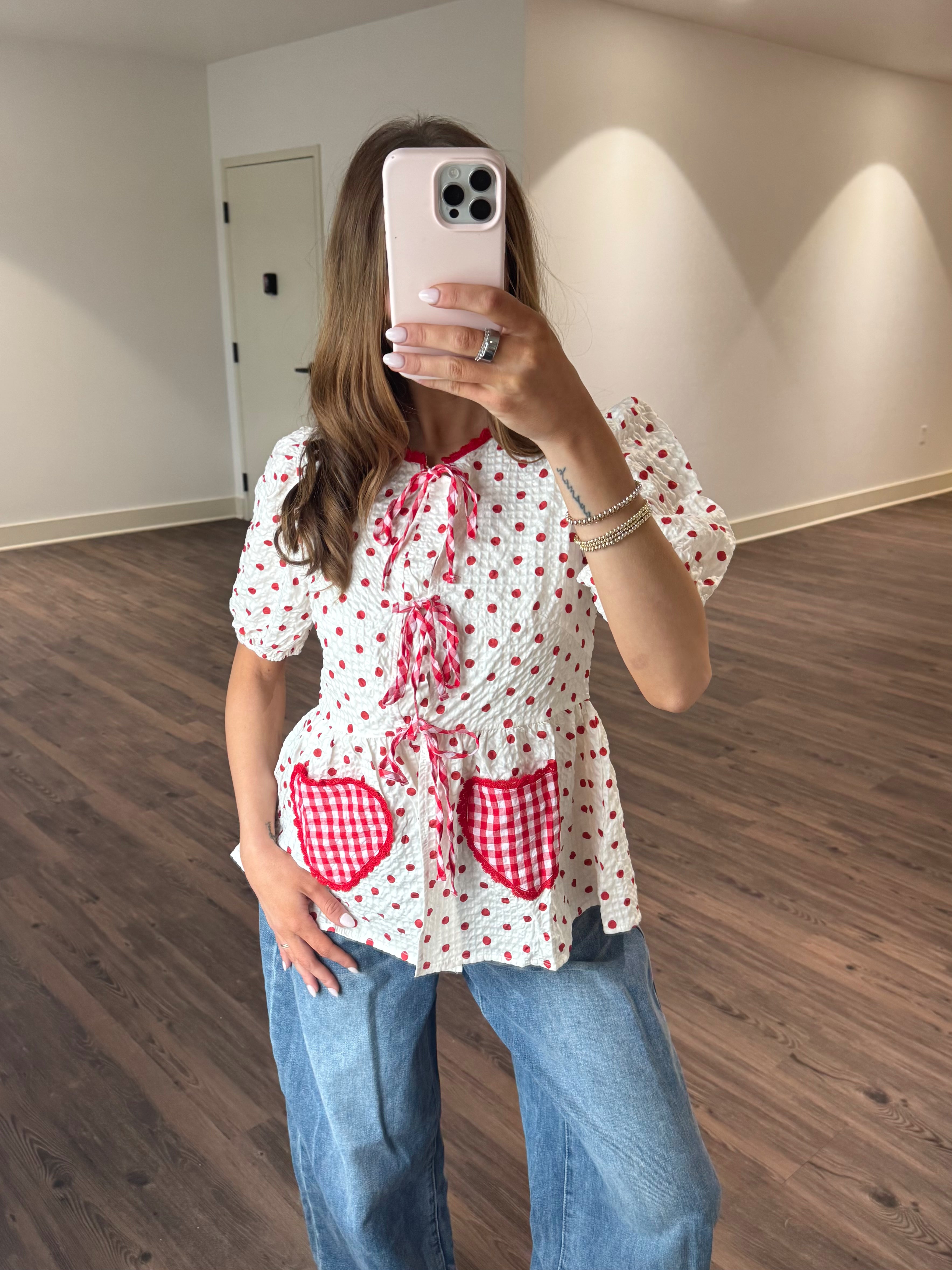Person wearing a white blouse with red heart patterns and blue jeans, taking a mirror selfie.