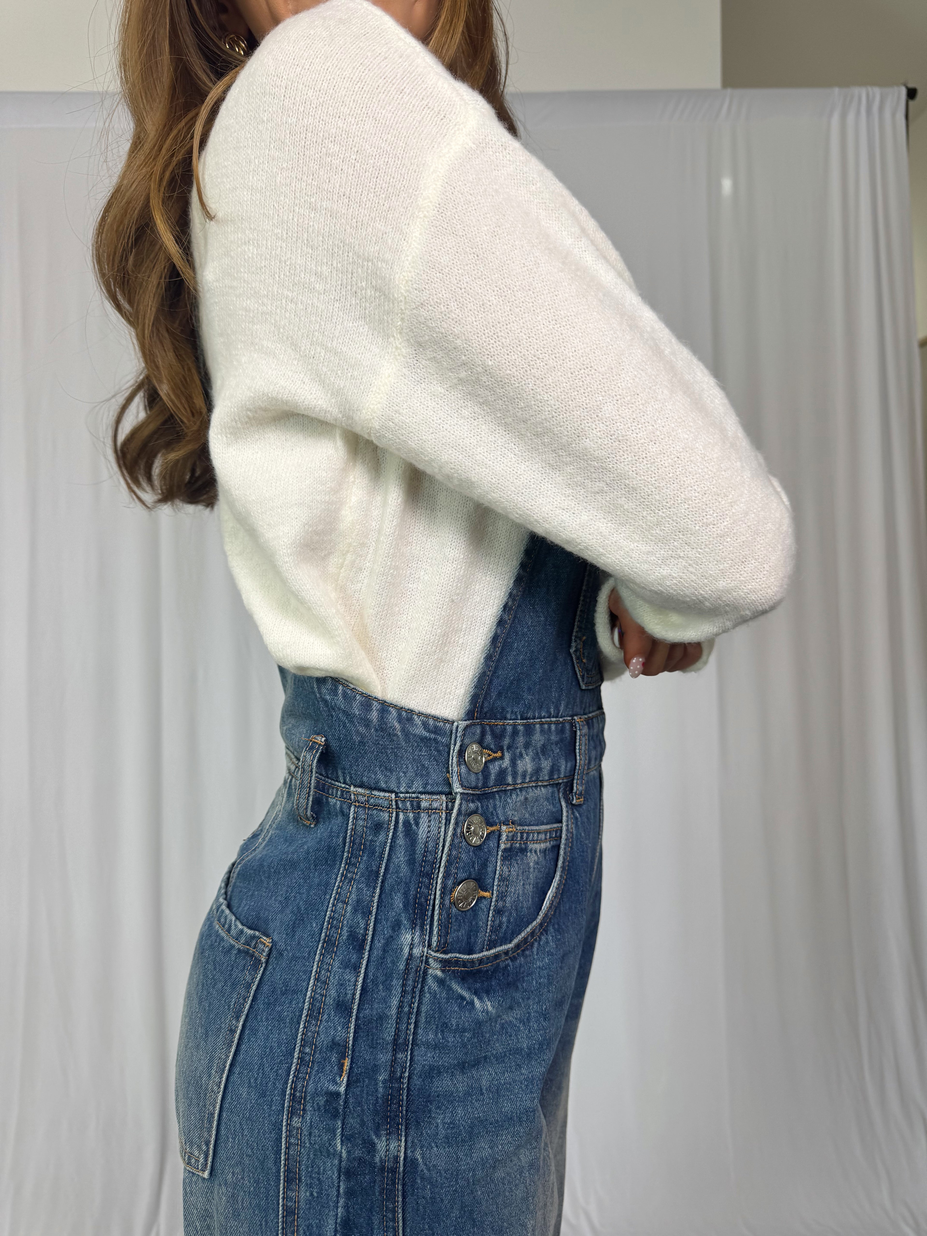 Person wearing a white sweater and blue jean overalls against a plain background