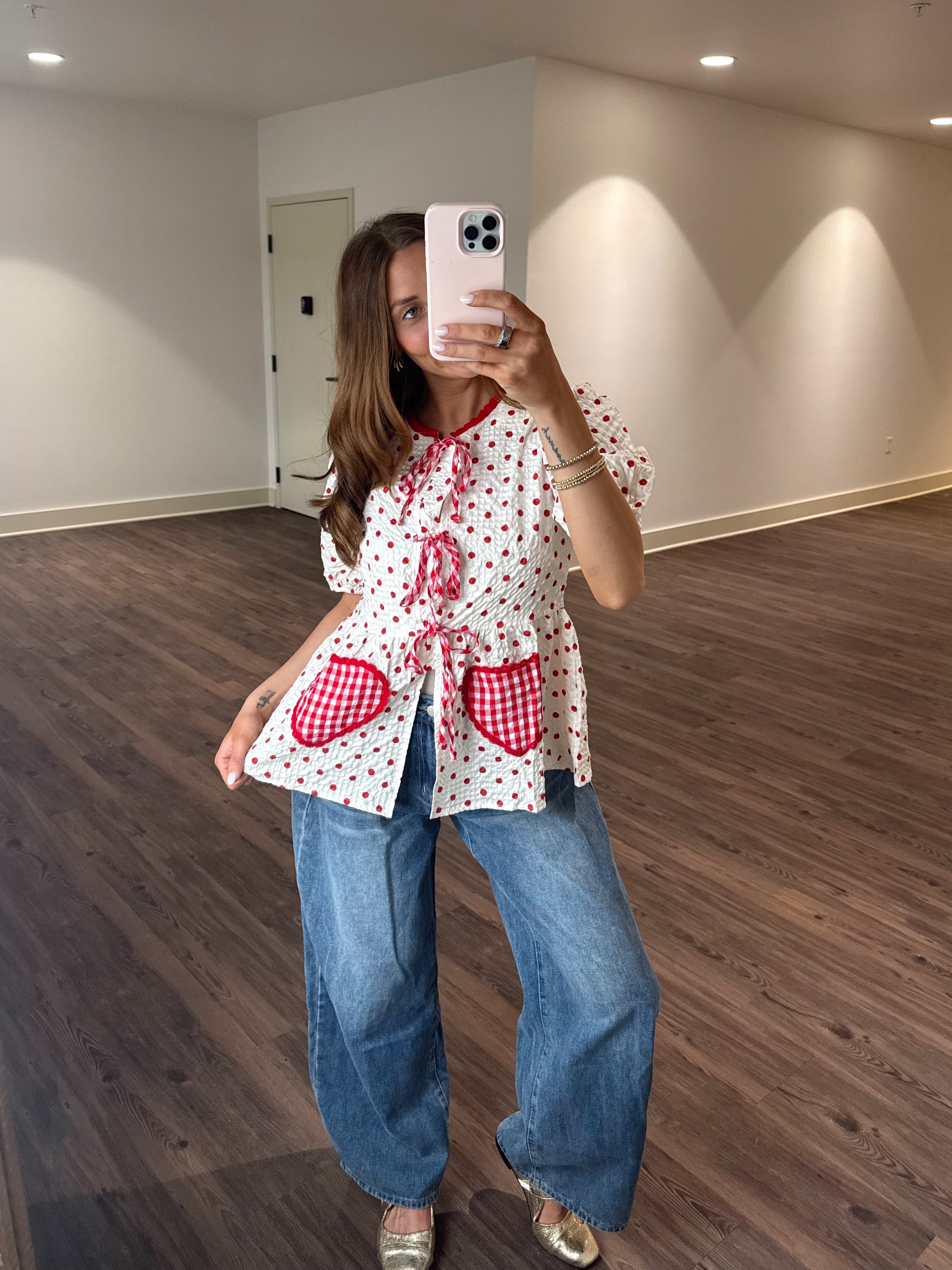 Person taking a mirror selfie wearing a white blouse with red patterns and blue jeans in a room with wooden flooring.