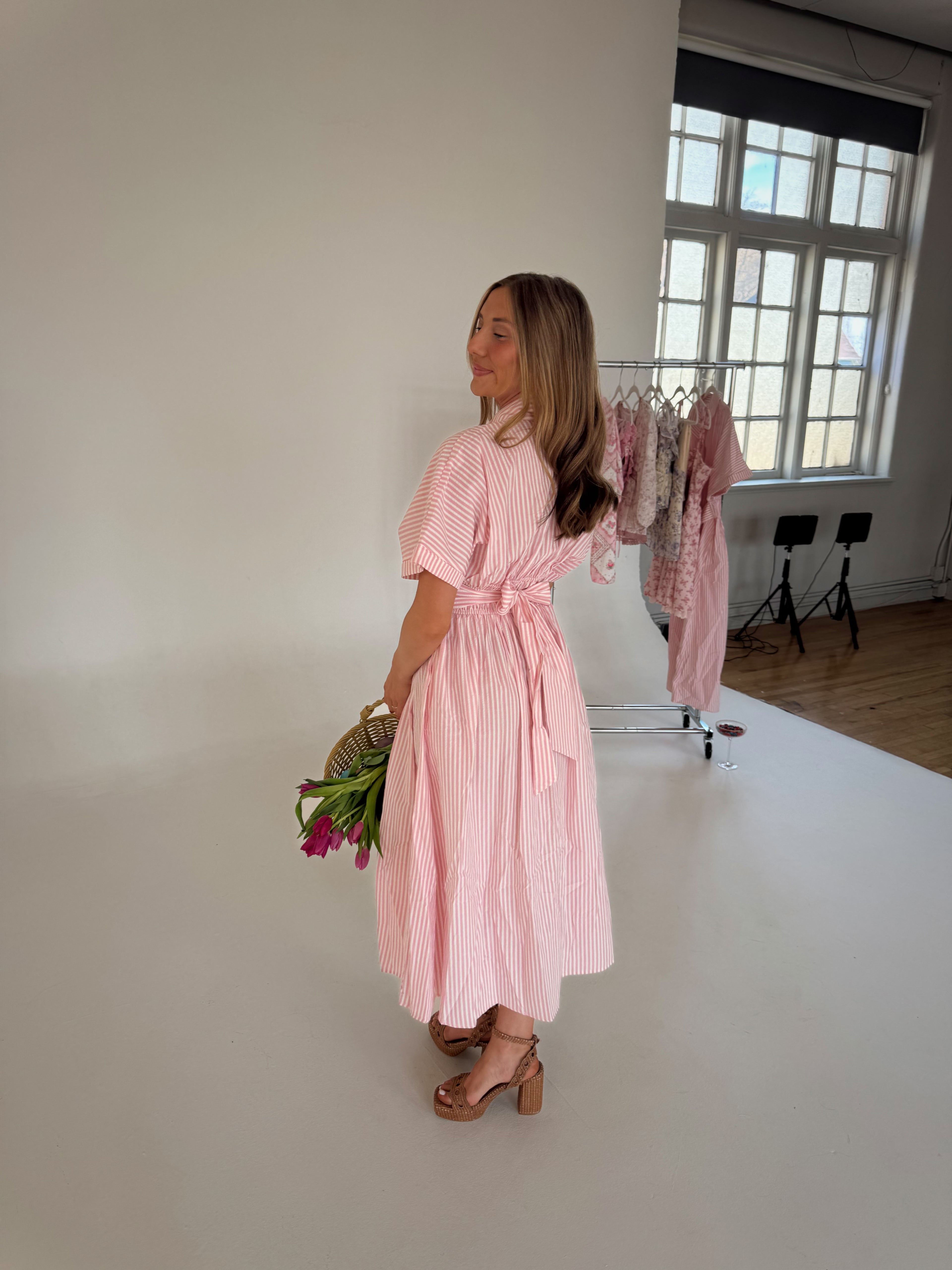 Woman in a pink dress holding flowers in a room with white walls and a window.