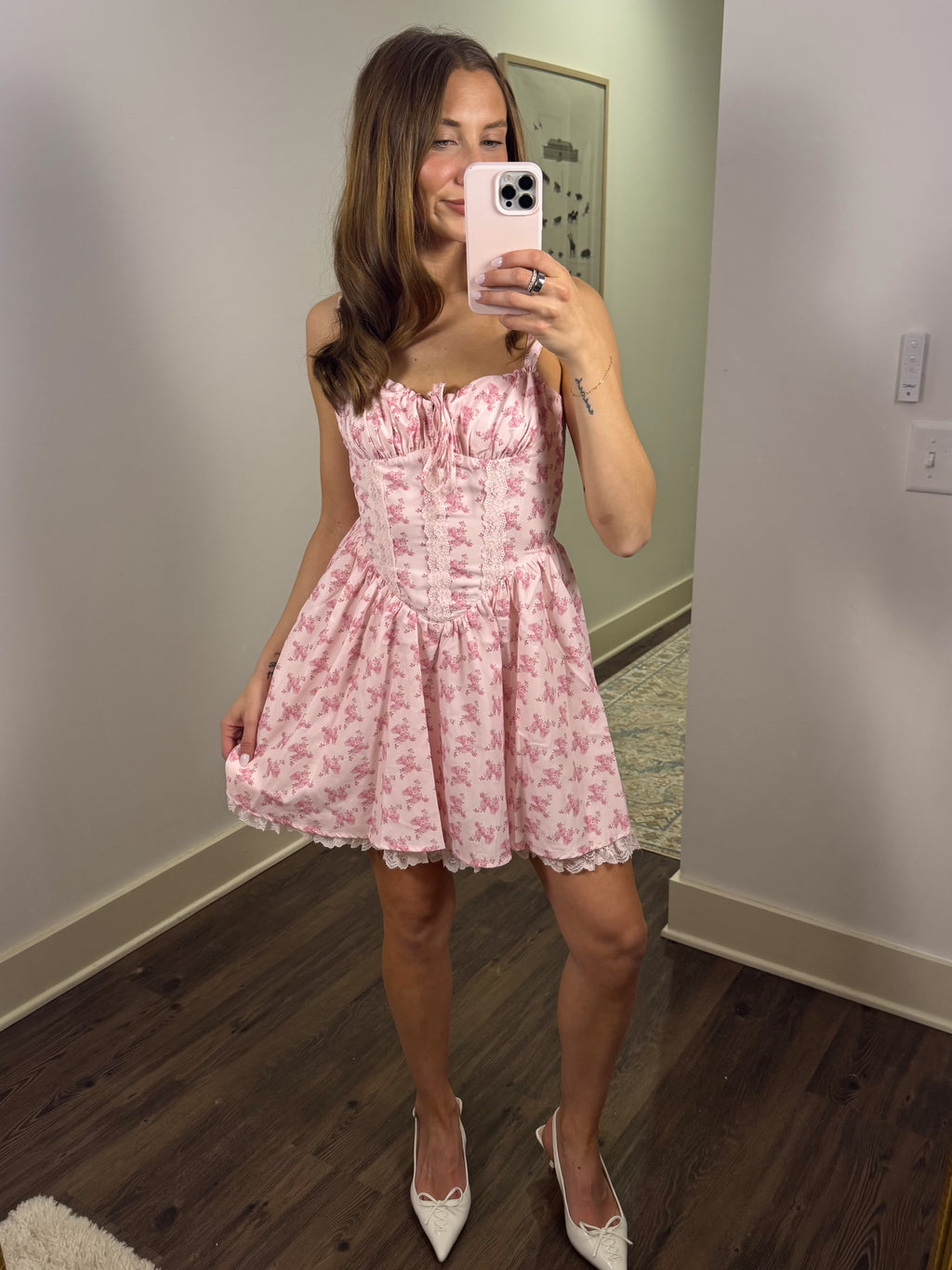 Woman taking a mirror selfie wearing a pink floral dress in a room with wooden flooring.