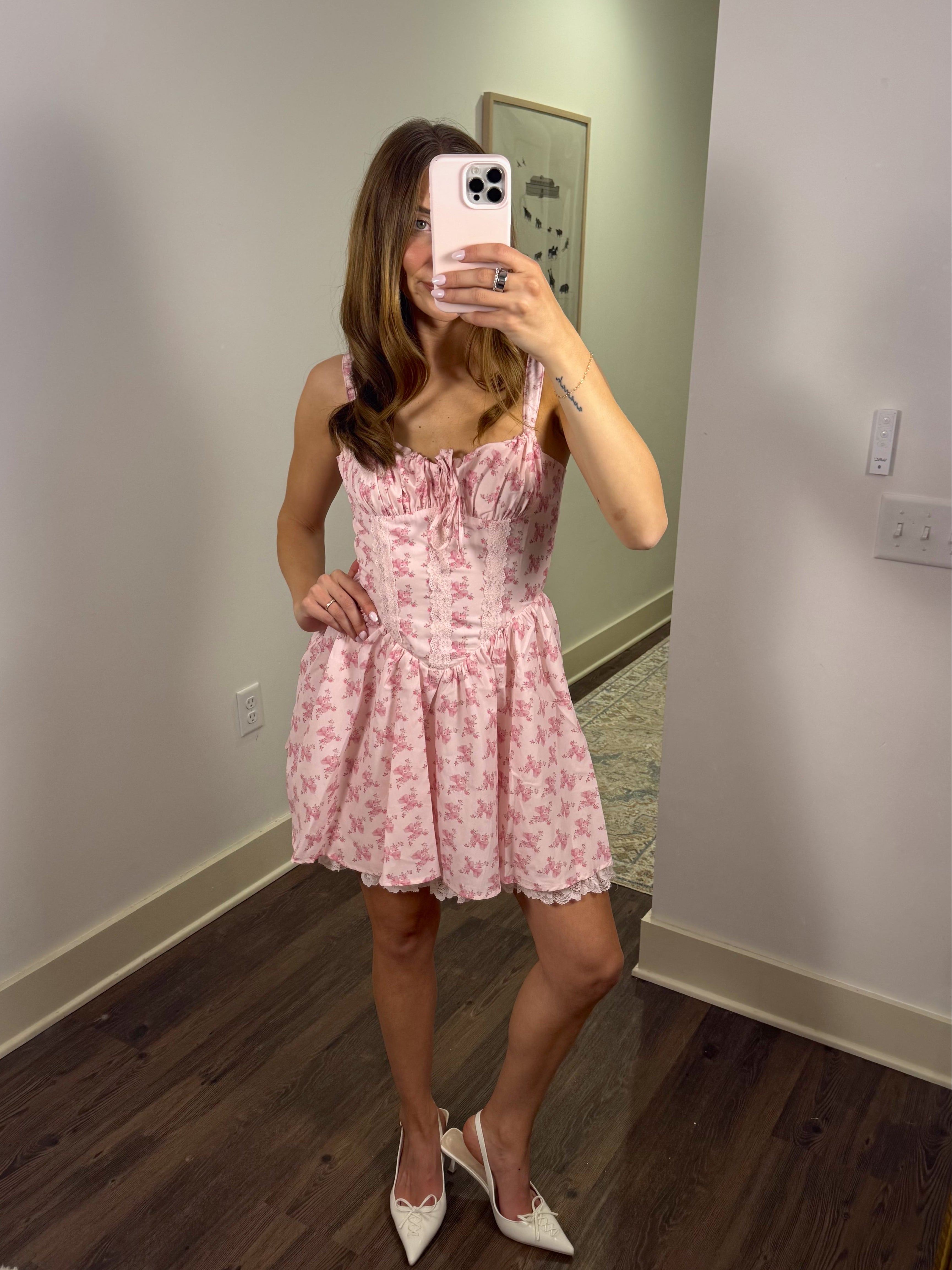 Woman taking a mirror selfie wearing a pink floral dress in a room with wooden flooring and a light gray wall.