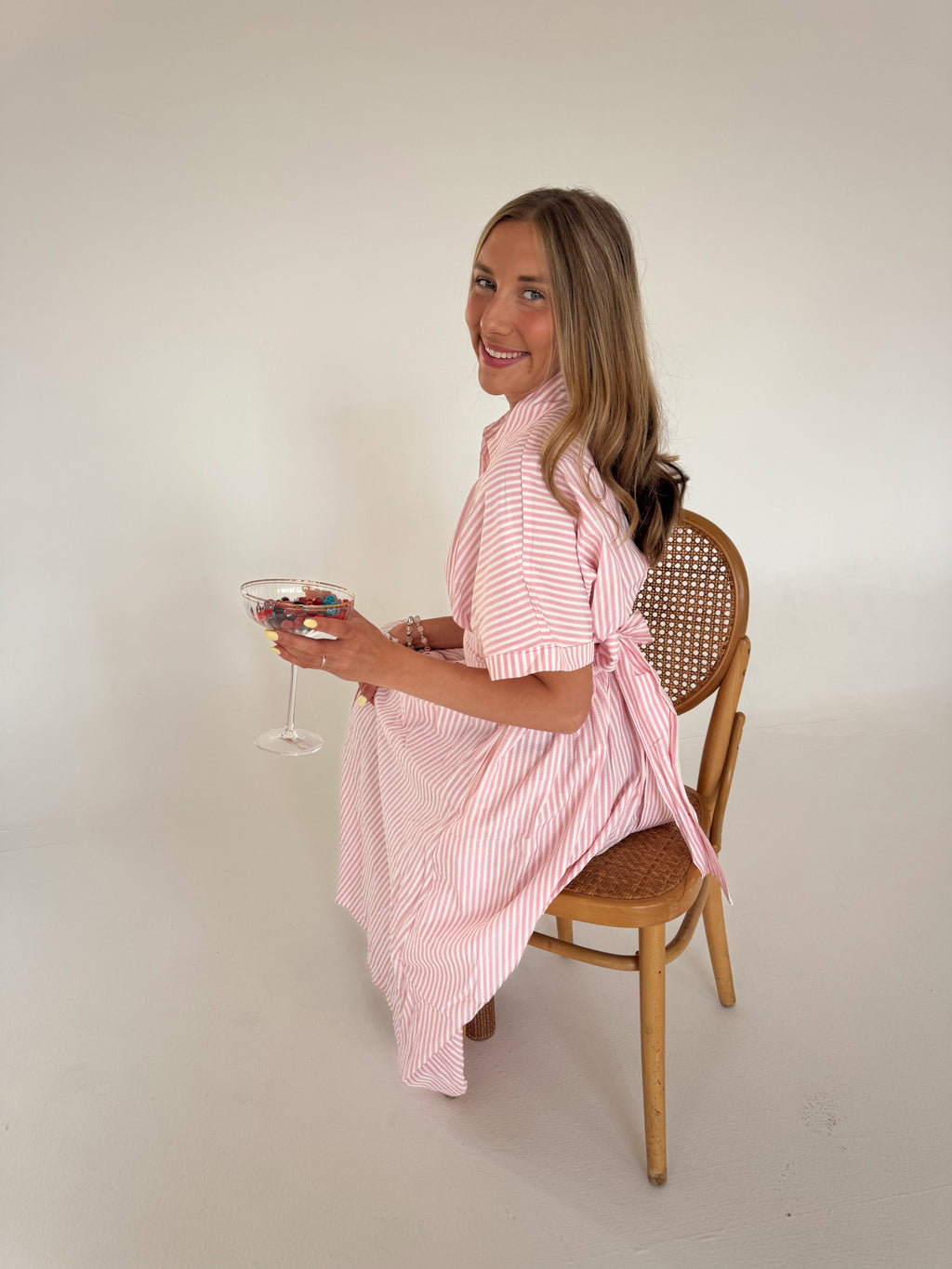 Woman in a pink and white striped dress sitting on a wooden chair holding a glass.