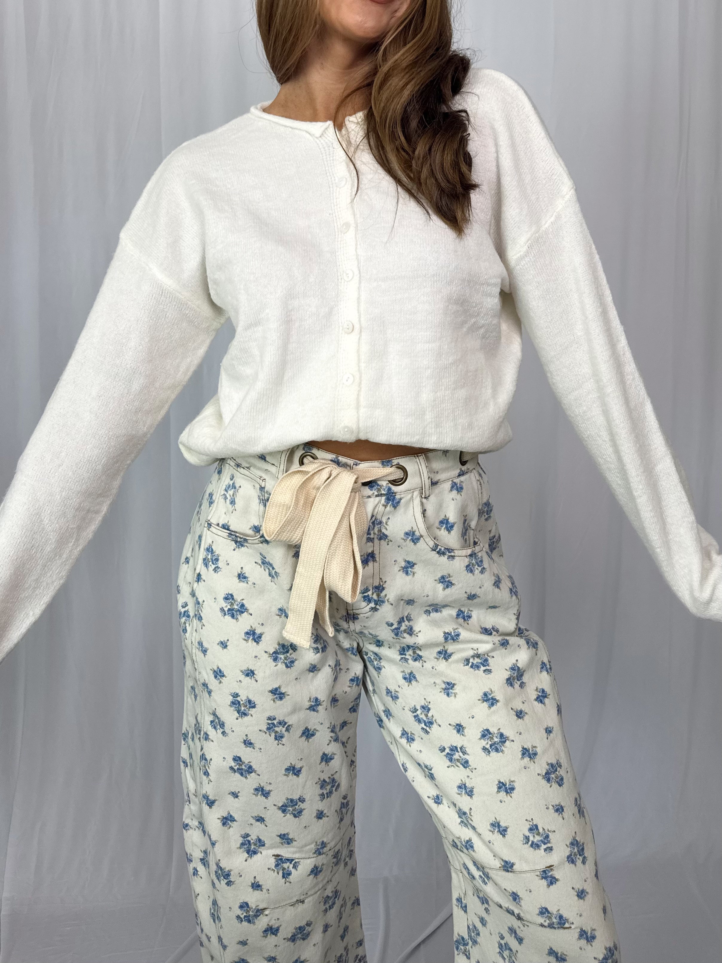 Woman wearing a white cardigan and floral pants against a plain background
