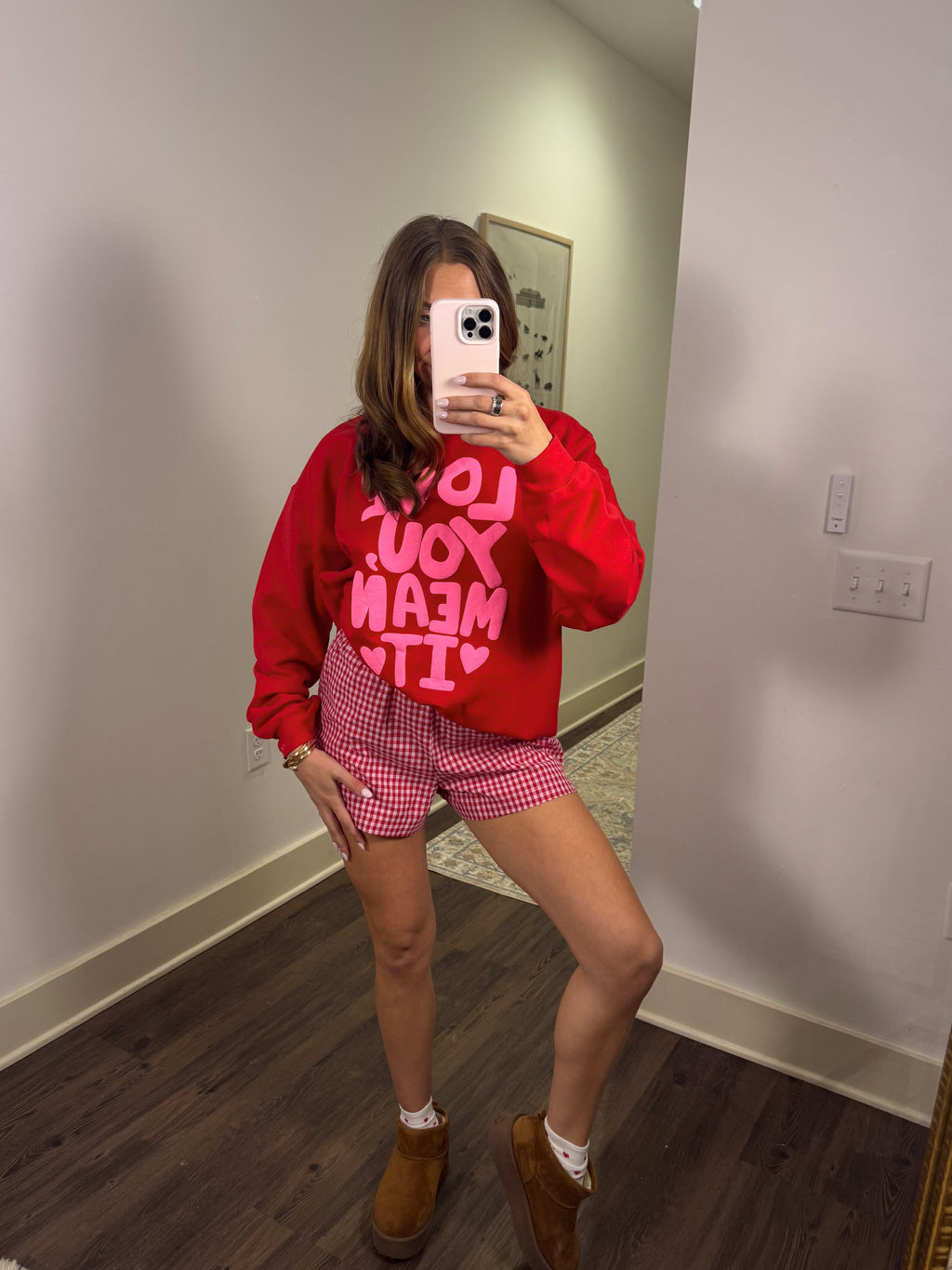 Person wearing a red sweatshirt with pink text and checkered shorts in a room.