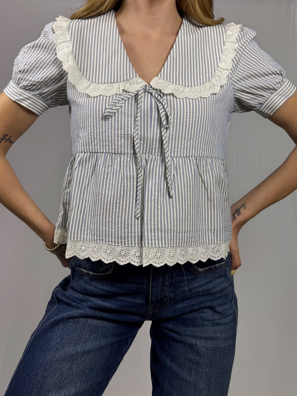 Woman wearing a blue and white striped blouse with lace details and blue jeans on a gray background