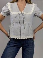 Woman wearing a blue and white striped blouse with lace details and blue jeans on a gray background