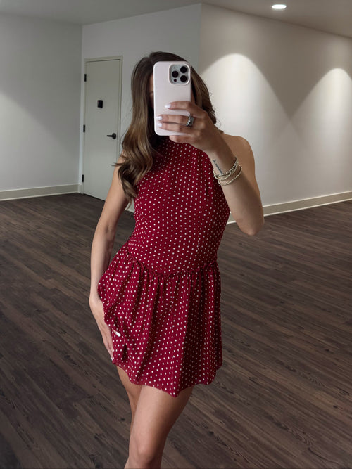 Woman taking a mirror selfie wearing a red polka dot dress in a room with wooden flooring and white walls.