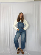 Person wearing blue denim overalls and a white long-sleeve shirt against a plain background