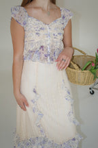 Woman wearing a floral skirt set holding a wicker basket with flowers against a plain background