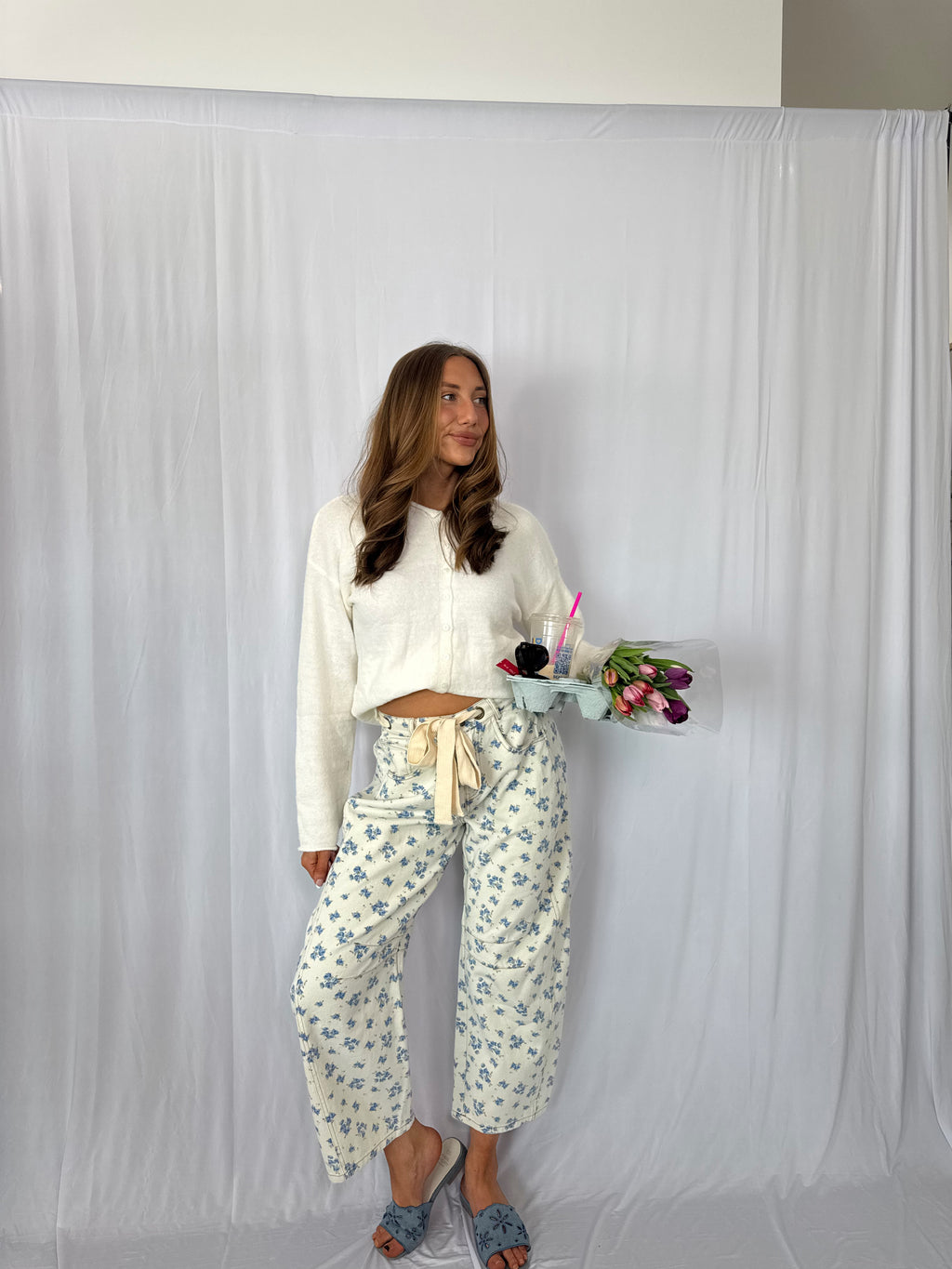 Woman in a white shirt and floral pants holding flowers against a plain backdrop