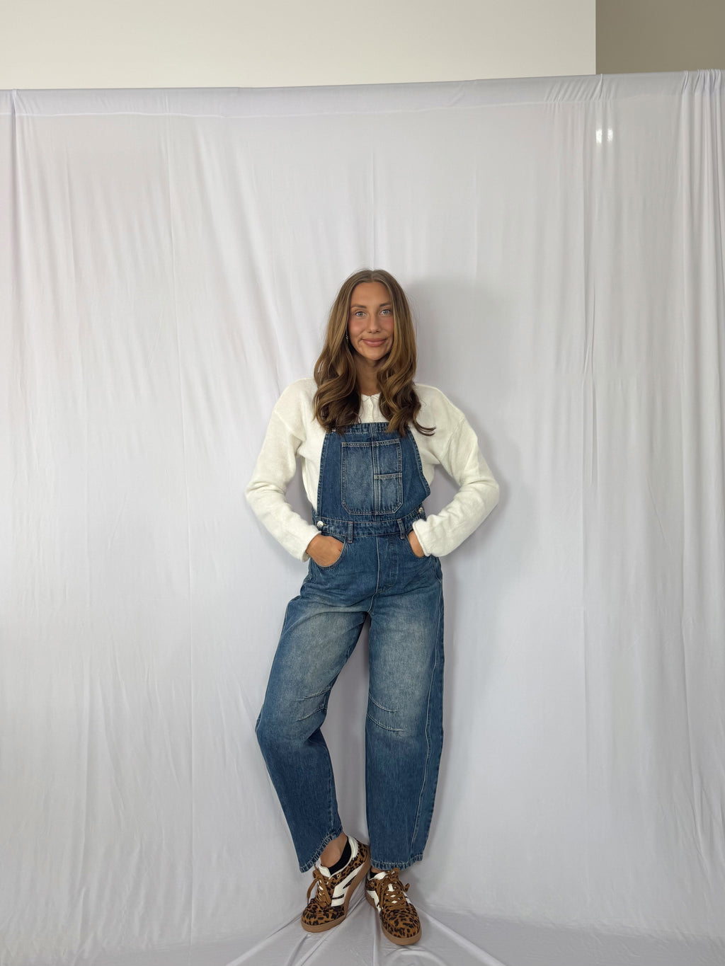 Person wearing blue denim overalls and a white long-sleeve shirt against a plain background