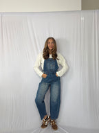 Person wearing blue denim overalls and a white long-sleeve shirt against a plain background