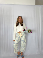 Woman in a white top and floral pants holding a tray with flowers and drinks against a plain background