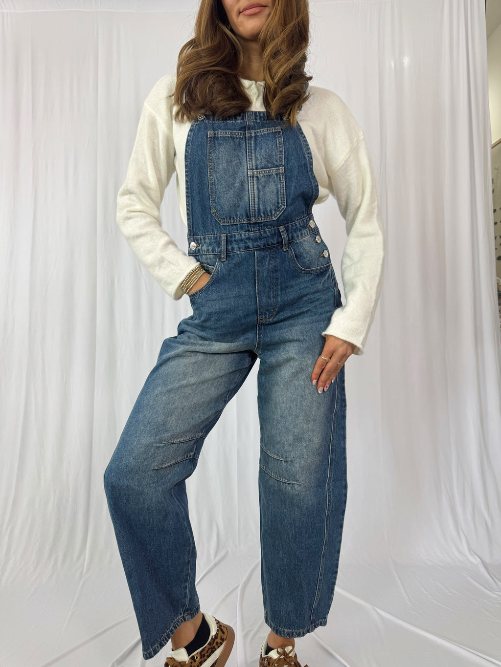 Person wearing blue denim overalls over a white long-sleeve shirt against a white curtain background