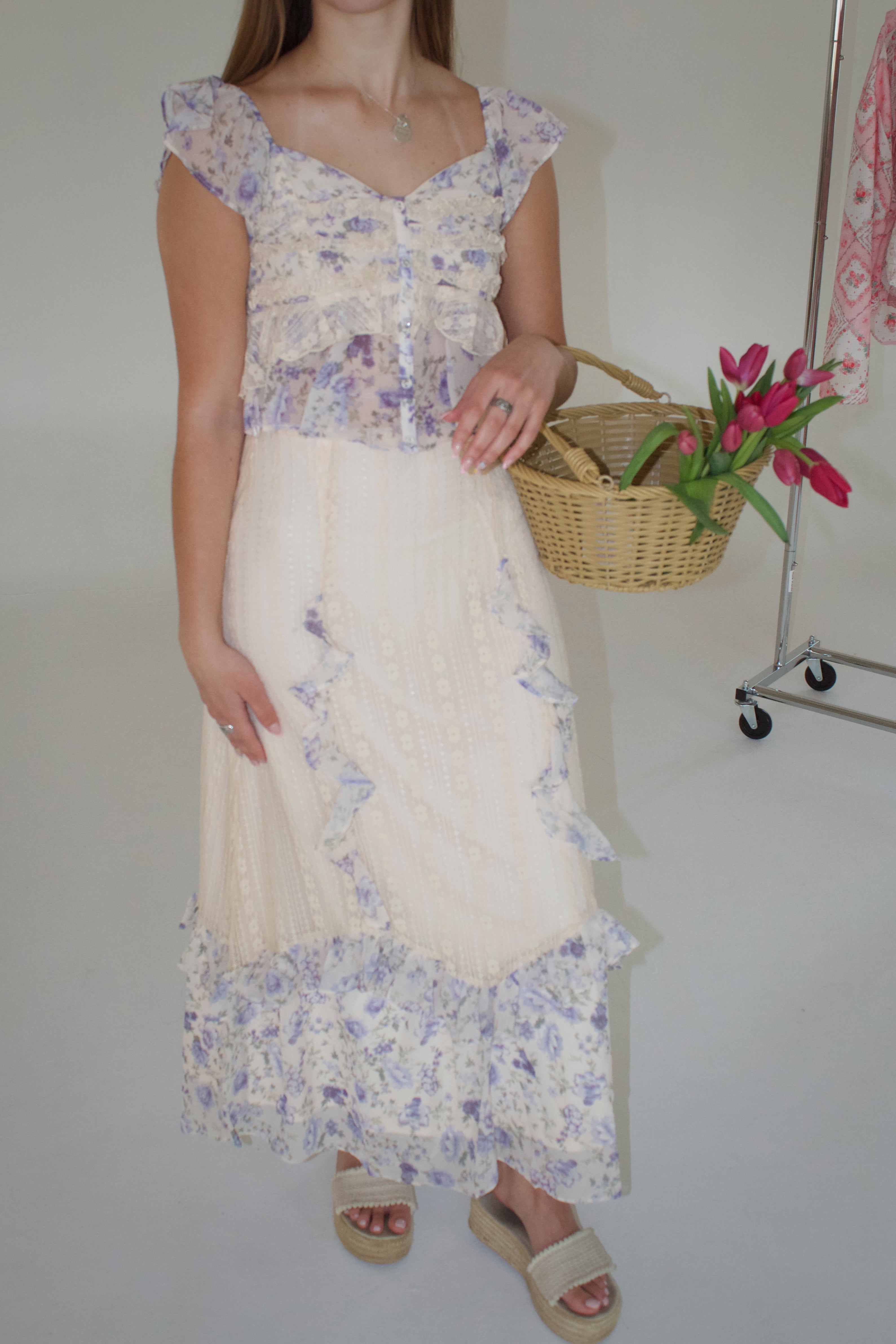Woman wearing a floral top and white skirt holding a basket of tulips against a plain background