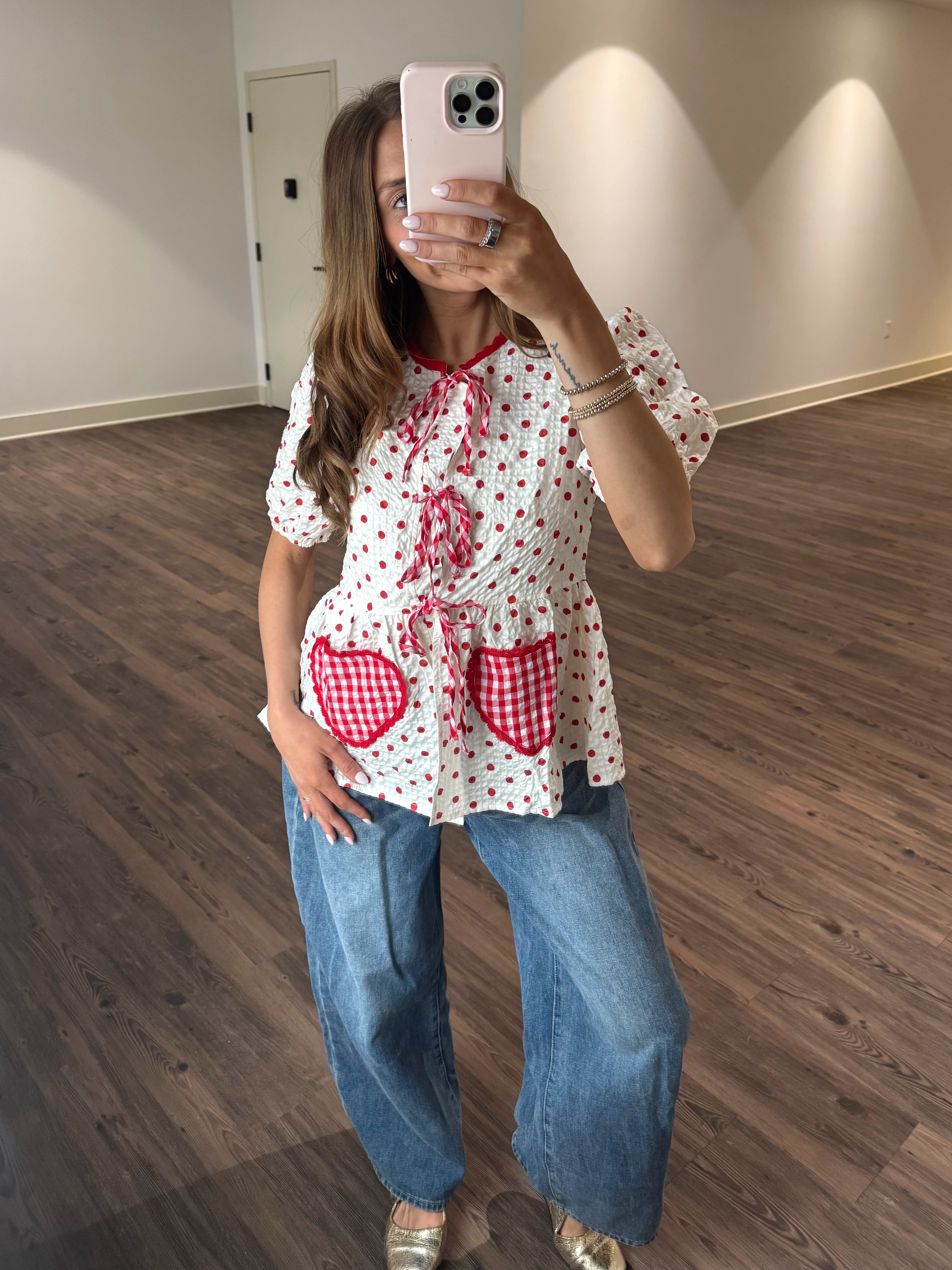 Person taking a mirror selfie wearing a polka dot blouse with red accents and blue jeans.