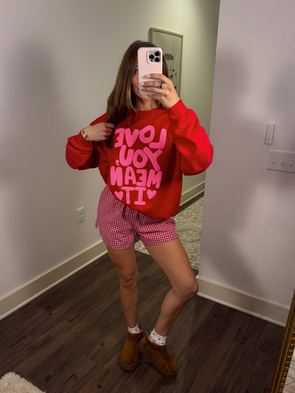 Person wearing a red sweatshirt with pink text and checkered shorts in a room.