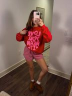 Person wearing a red sweatshirt with pink text and checkered shorts in a room.