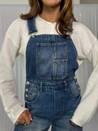 Person wearing blue denim overalls over a white long-sleeve shirt against a neutral background