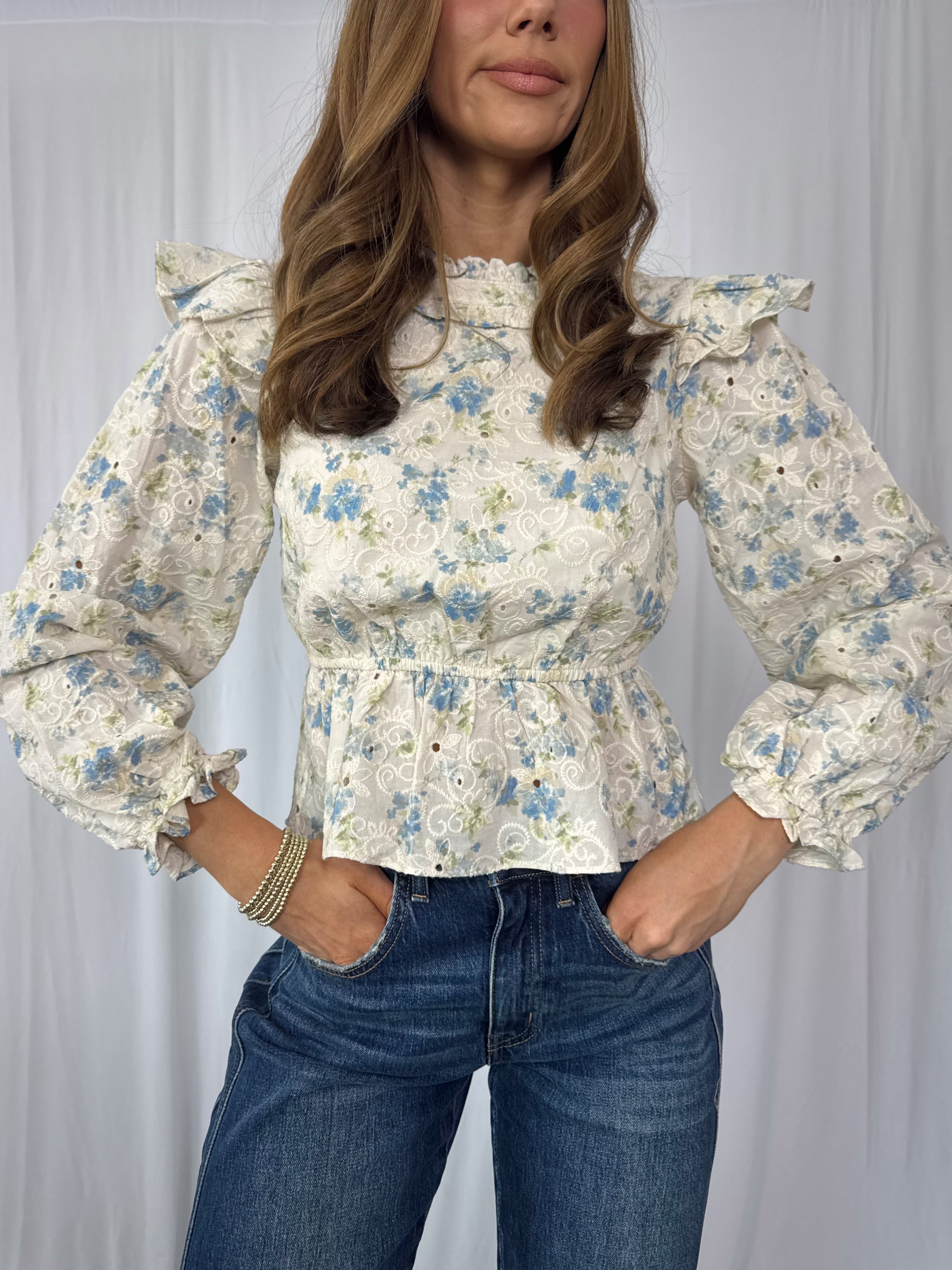 Woman wearing a floral blouse with long sleeves and blue jeans against a plain background