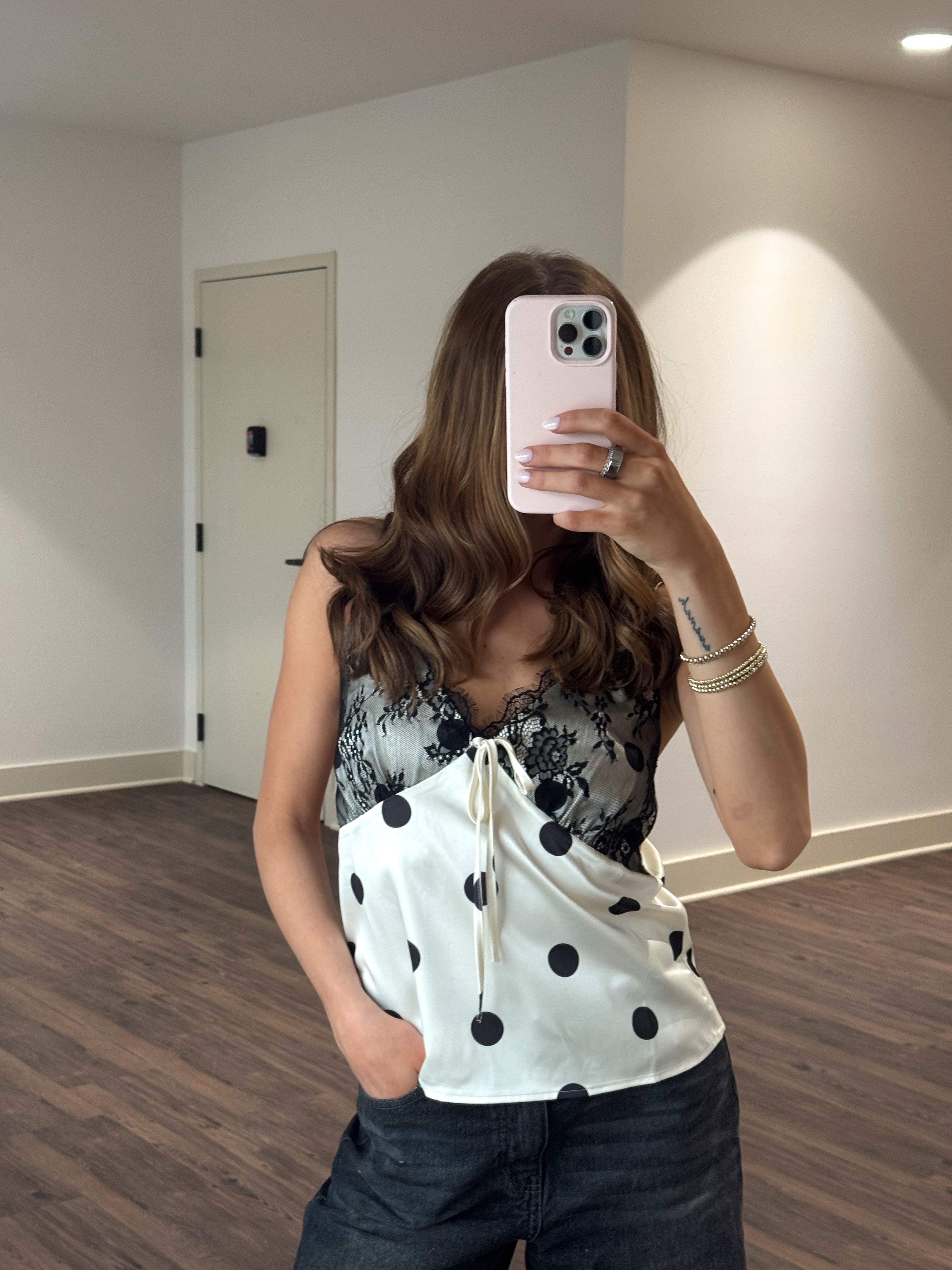 Person taking a mirror selfie wearing a polka dot top and black pants in a room with wooden flooring.