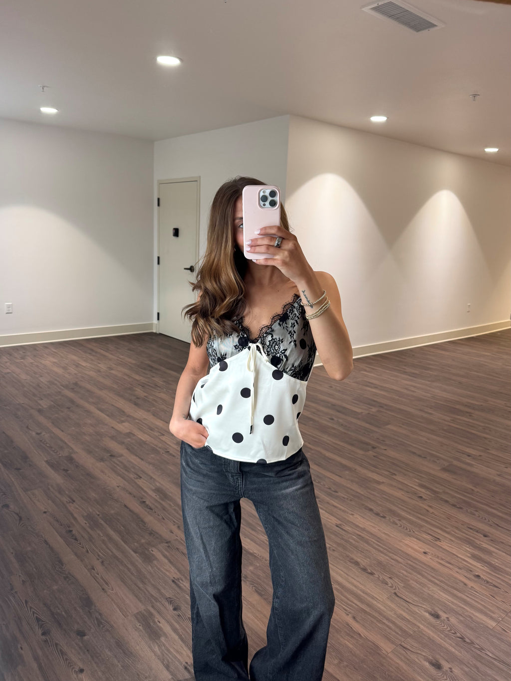 Woman taking a mirror selfie of a white top with black polka dots in a room with wooden flooring and white walls.