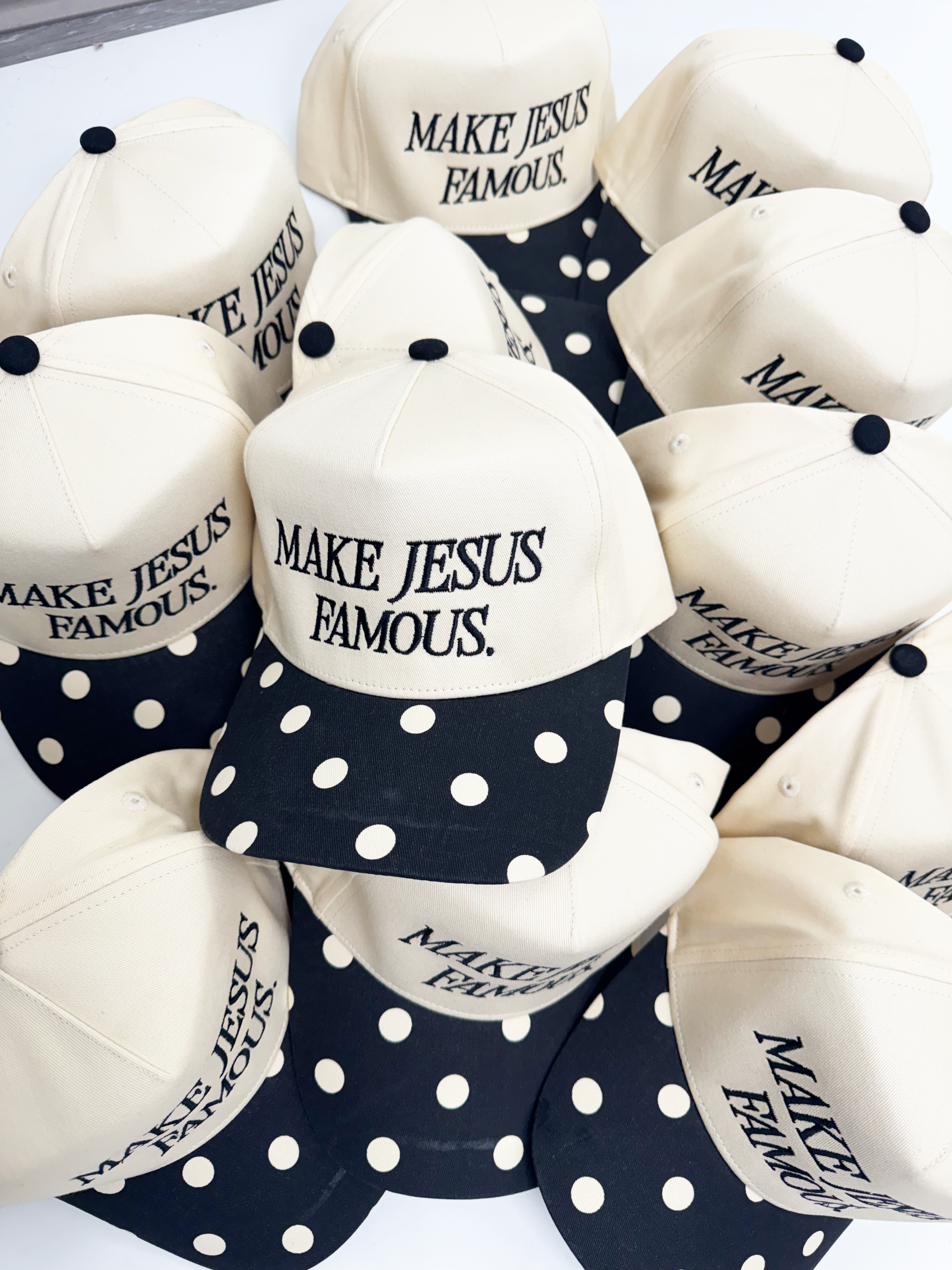 Collection of hats with 'MAKE JESUS FAMOUS' text in black embroidered on a white background. The bill of the hat is black with white polka dots.