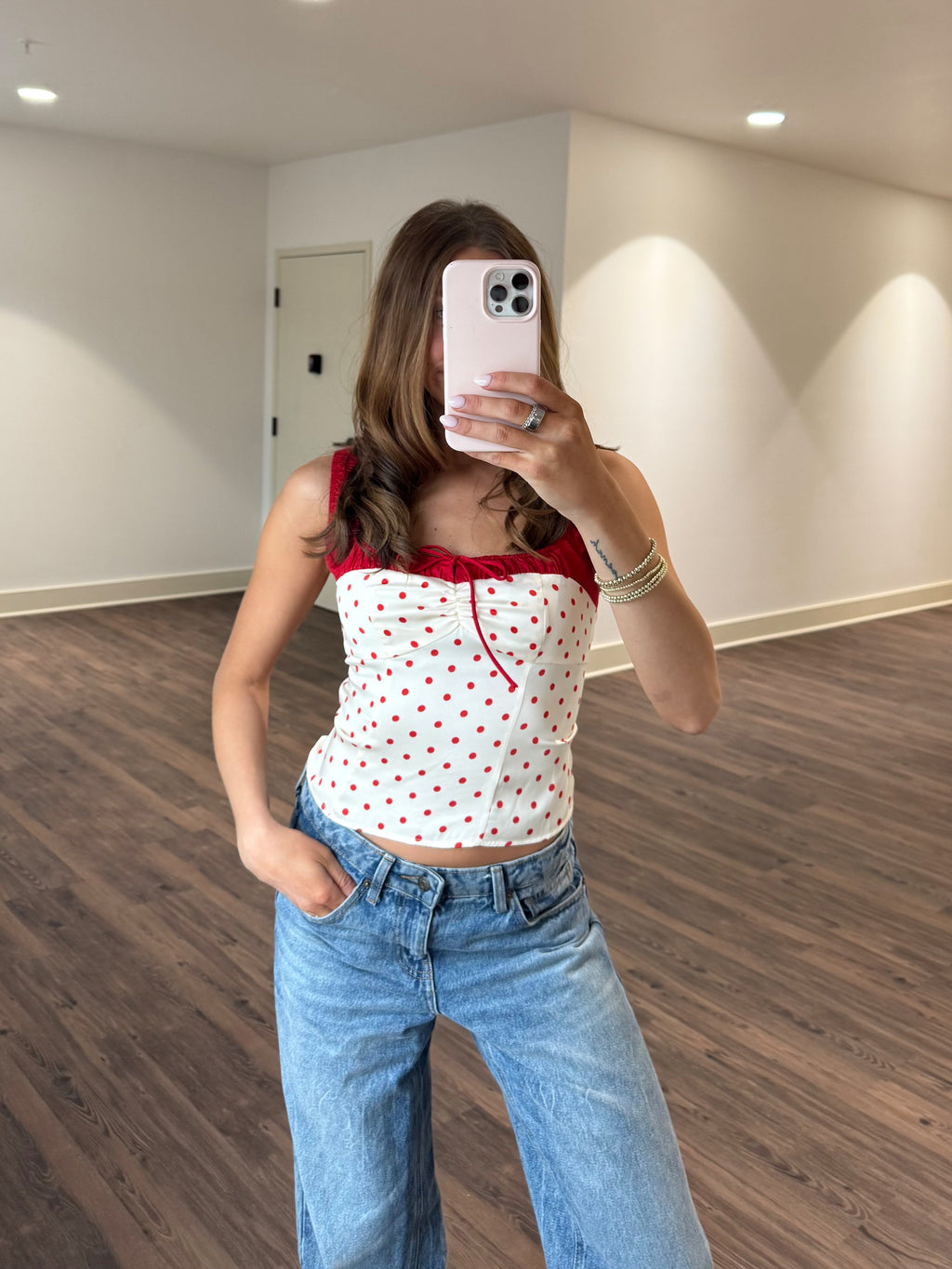 Person taking a mirror selfie wearing a white top with red polka dots and blue jeans in a room with wooden flooring and white walls.