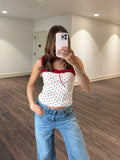 Person taking a mirror selfie wearing a white top with red polka dots and blue jeans in a room with wooden flooring and white walls.