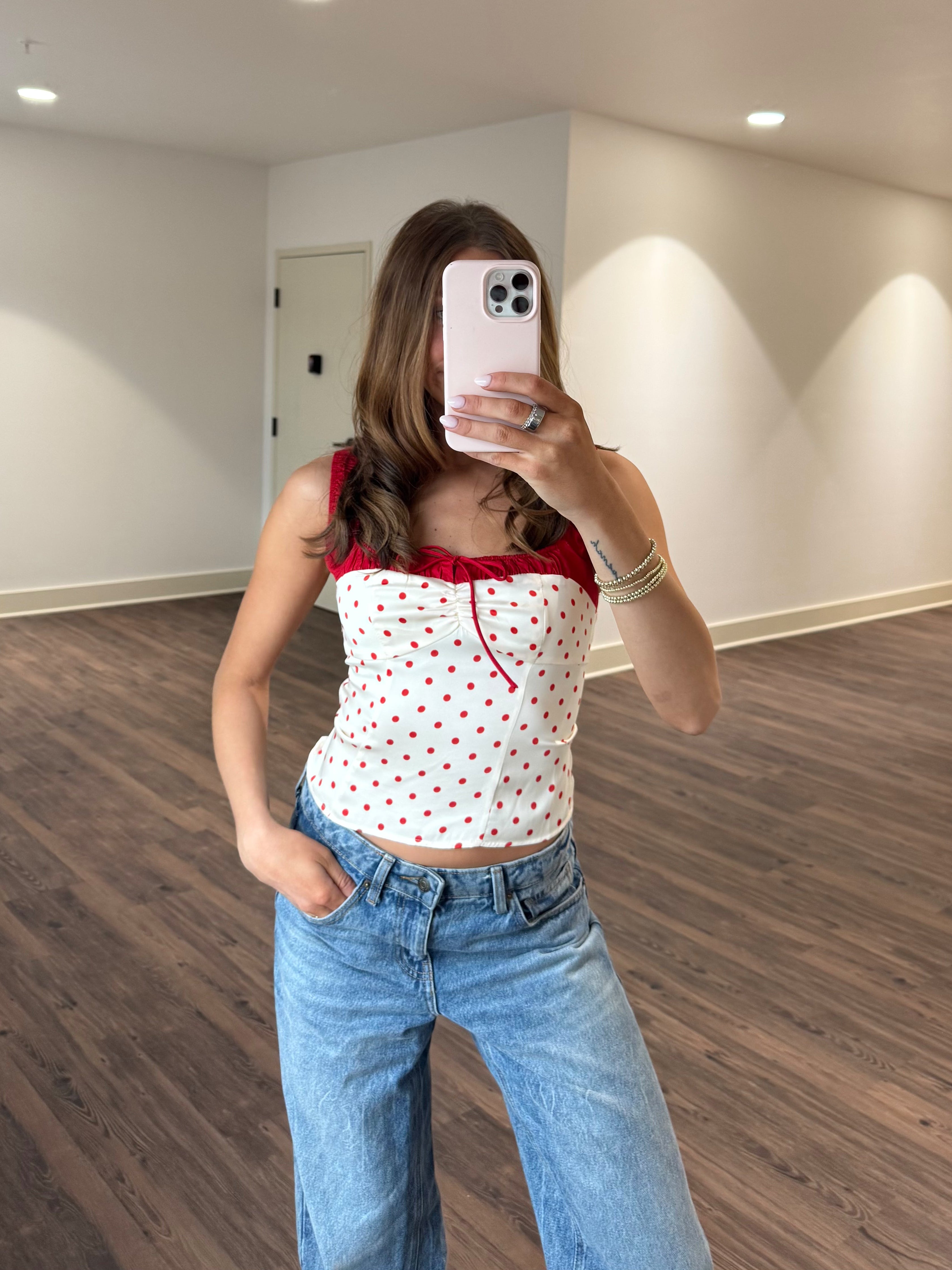 Person taking a mirror selfie wearing a white top with red polka dots and blue jeans in a room with wooden flooring and white walls.