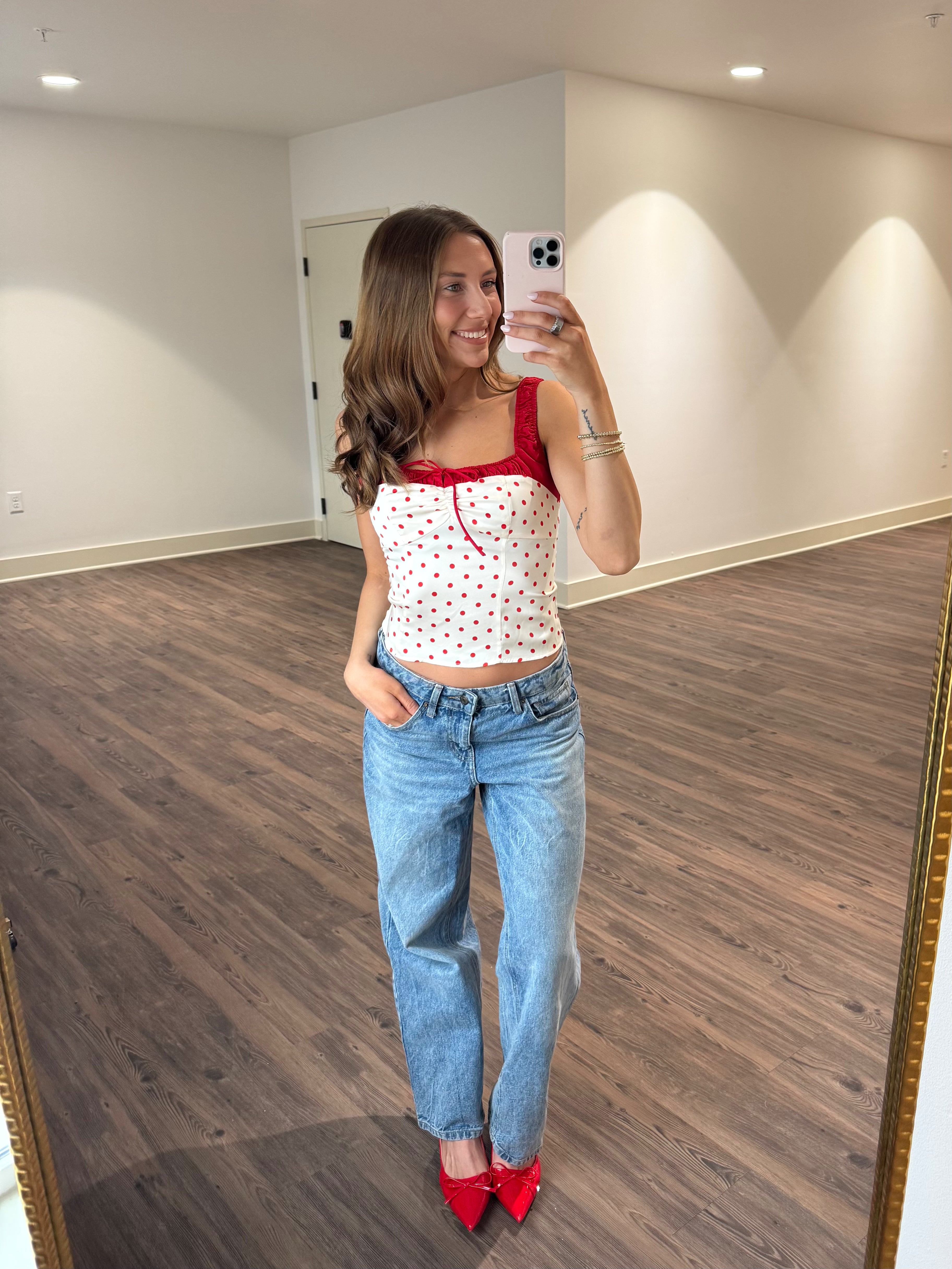 Woman taking a mirror selfie wearing a red top, white polka dot crop top, blue jeans, and red shoes in a room with wooden flooring.