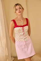 Woman wearing a white top with red polka dots and a pink skirt against a beige curtain background