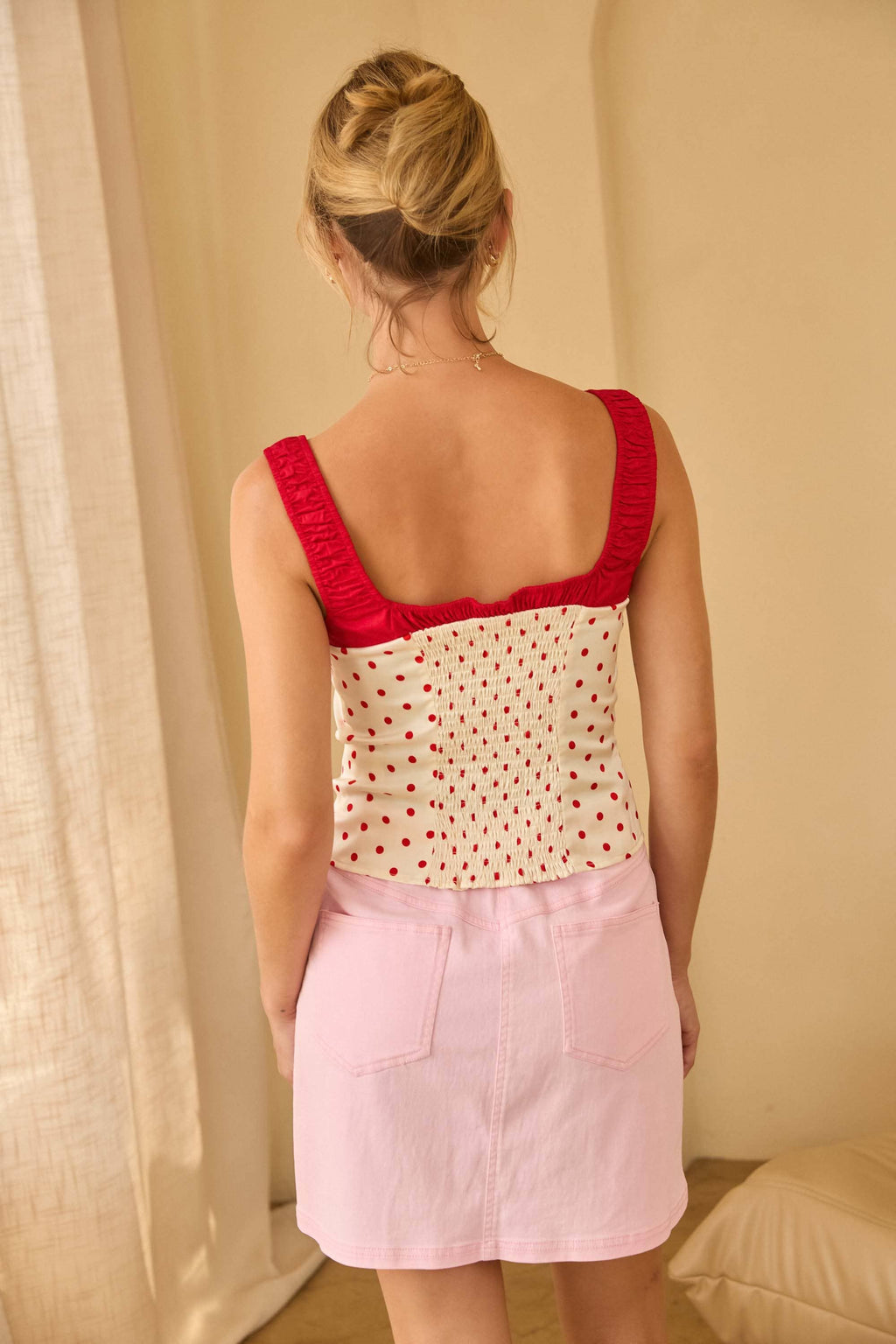 Woman wearing a red top and pink skirt standing in a room with beige walls and curtains. She is showing the back of the shirt.