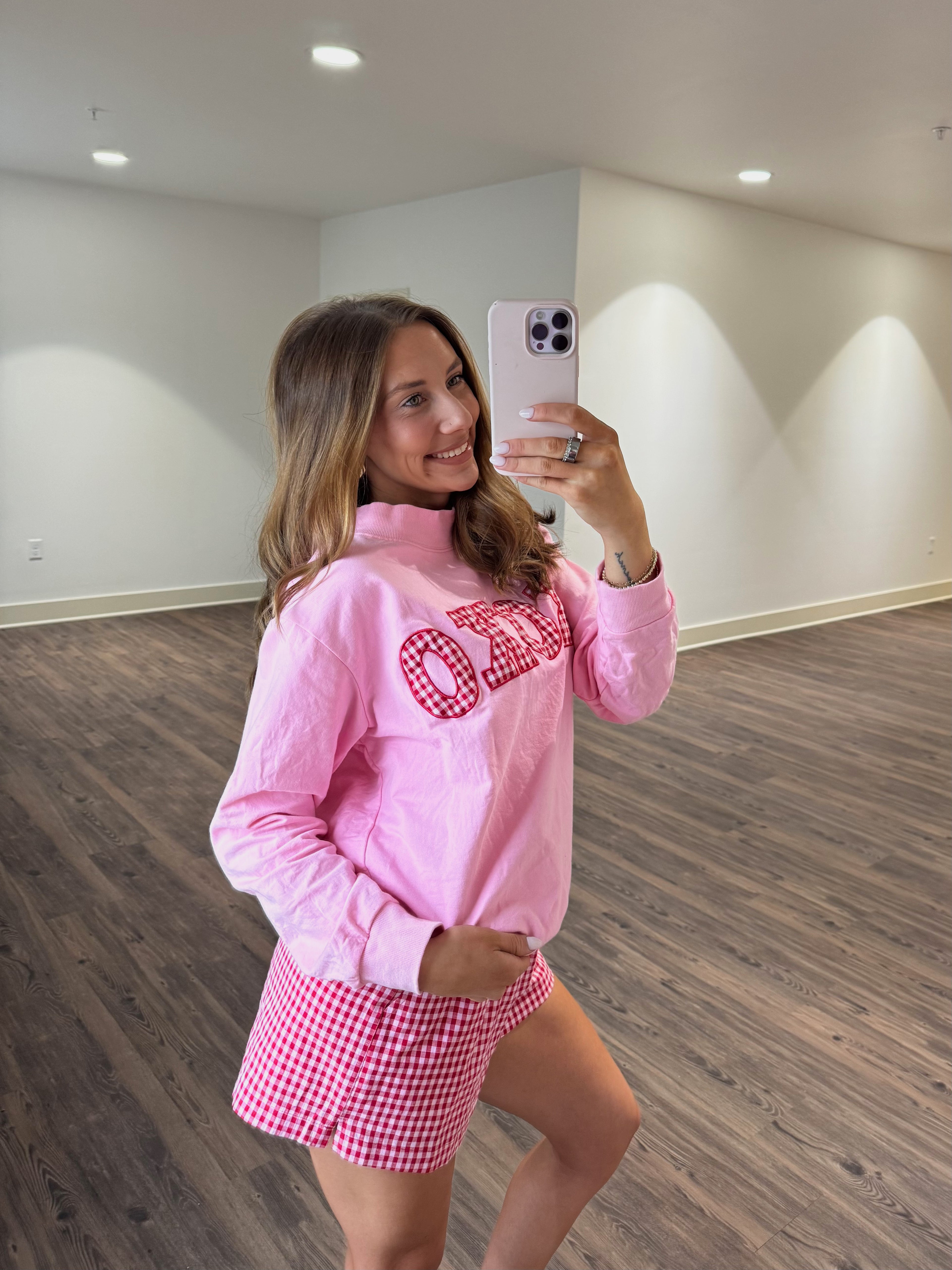 Woman taking a mirror selfie wearing a pink sweatshirt with text and red checkered shorts.