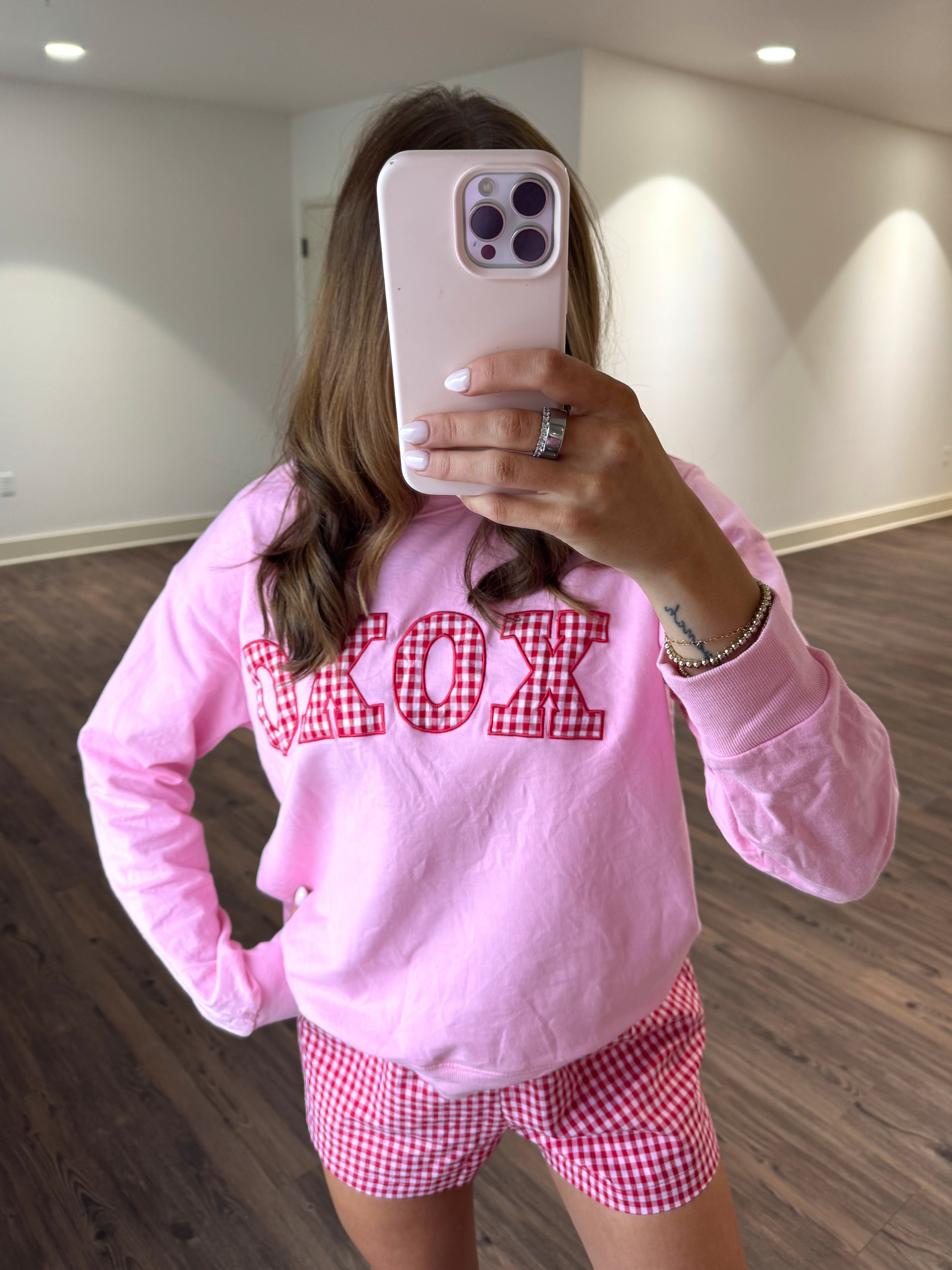 Person wearing a pink sweatshirt with 'XOXO' and red checkered shorts in a room.